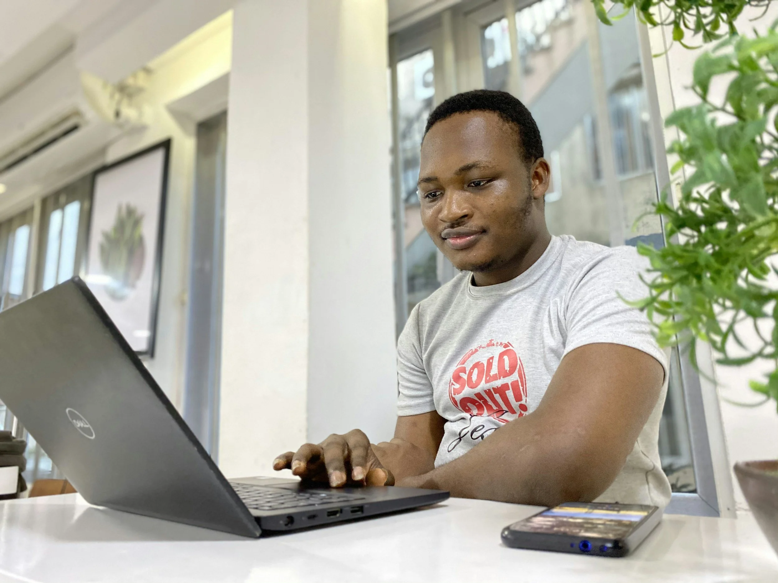 A young man wearing a gray t-shirt with a red and white graphic, sitting at a white table and working on a black Dell laptop in a bright room with large windows. A smartphone is next to his right arm and a green plant is partially visible on the right side of the image.