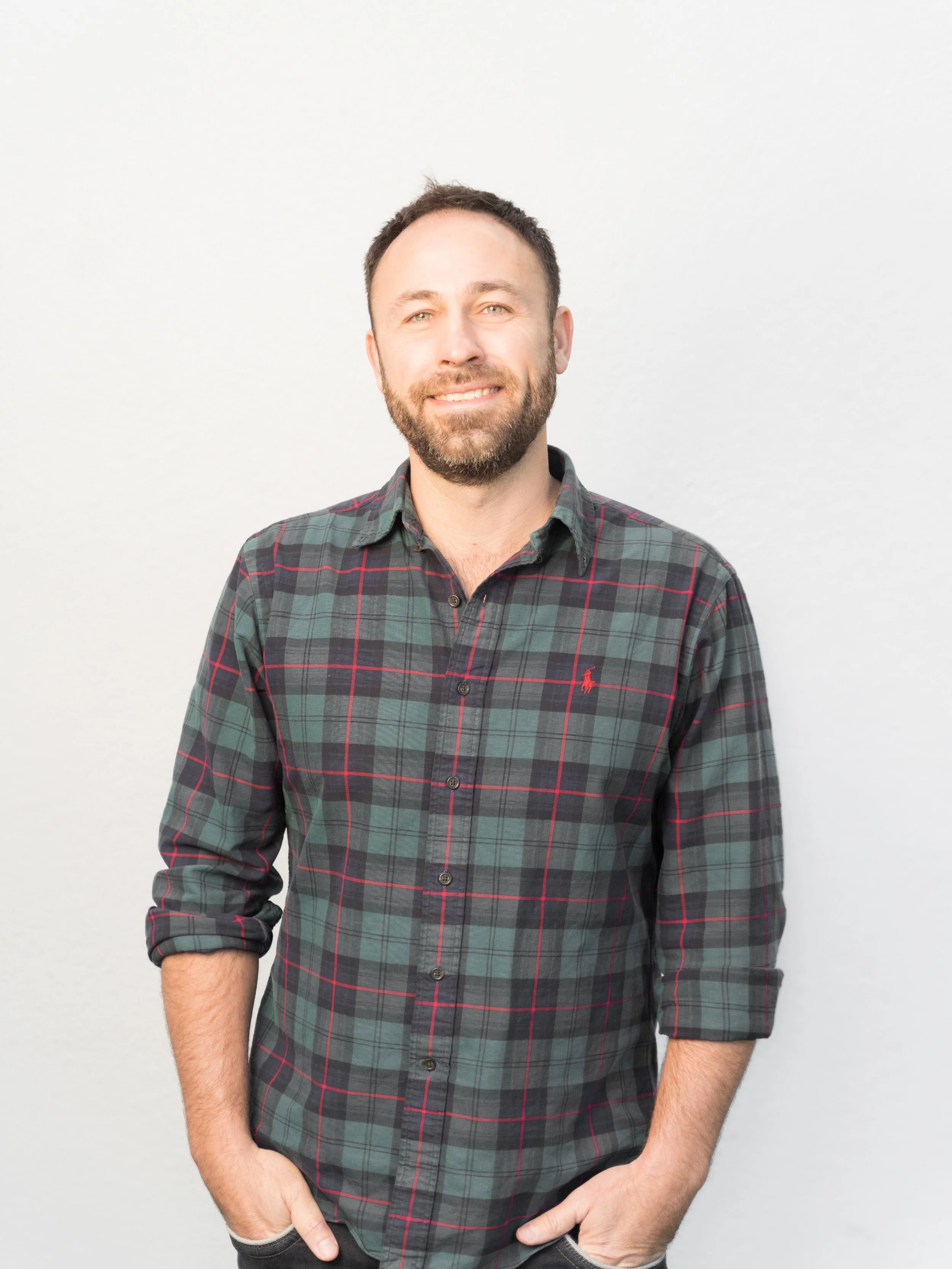 A man with a beard and short dark hair smiling, wearing a gray and red plaid button-up shirt with rolled-up sleeves, standing against a plain light background.