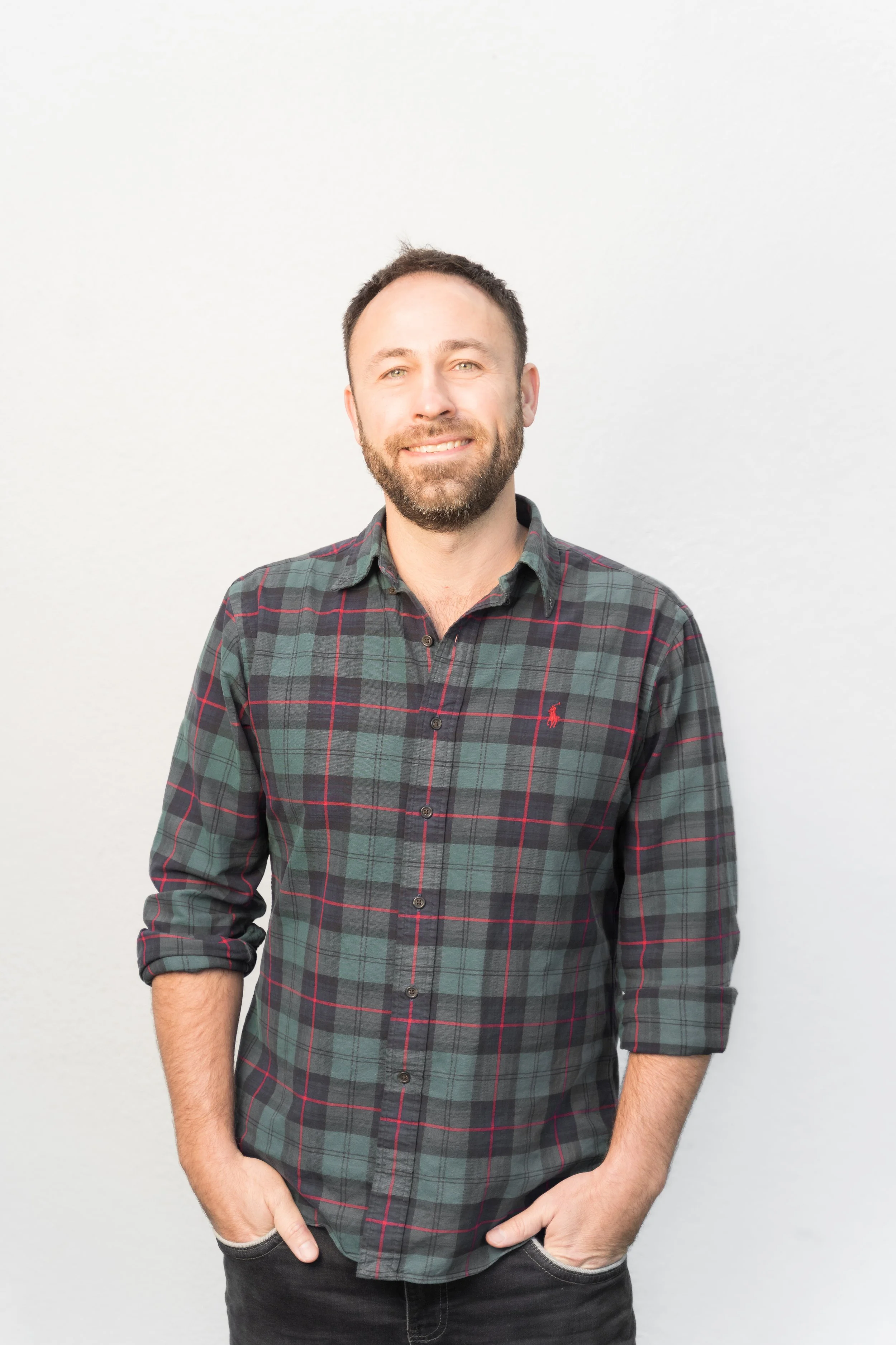 A smiling man with a beard and short hair, wearing a green and red plaid shirt with rolled-up sleeves, standing against a plain white background.