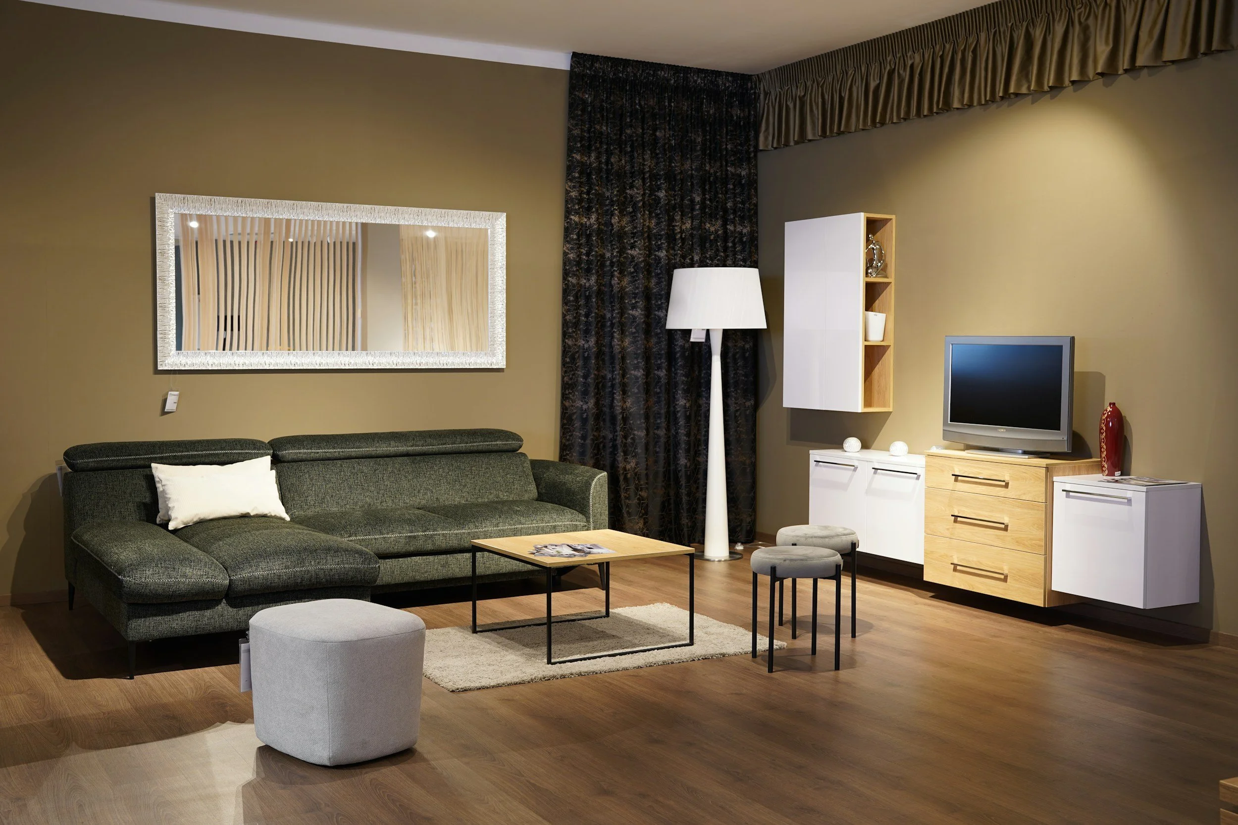 Living room with green sofa, wooden coffee table, gray ottoman, wall mirror, standing white lamp, wall-mounted cabinet, TV on a wooden stand, decorative items, and dark curtains on the window.