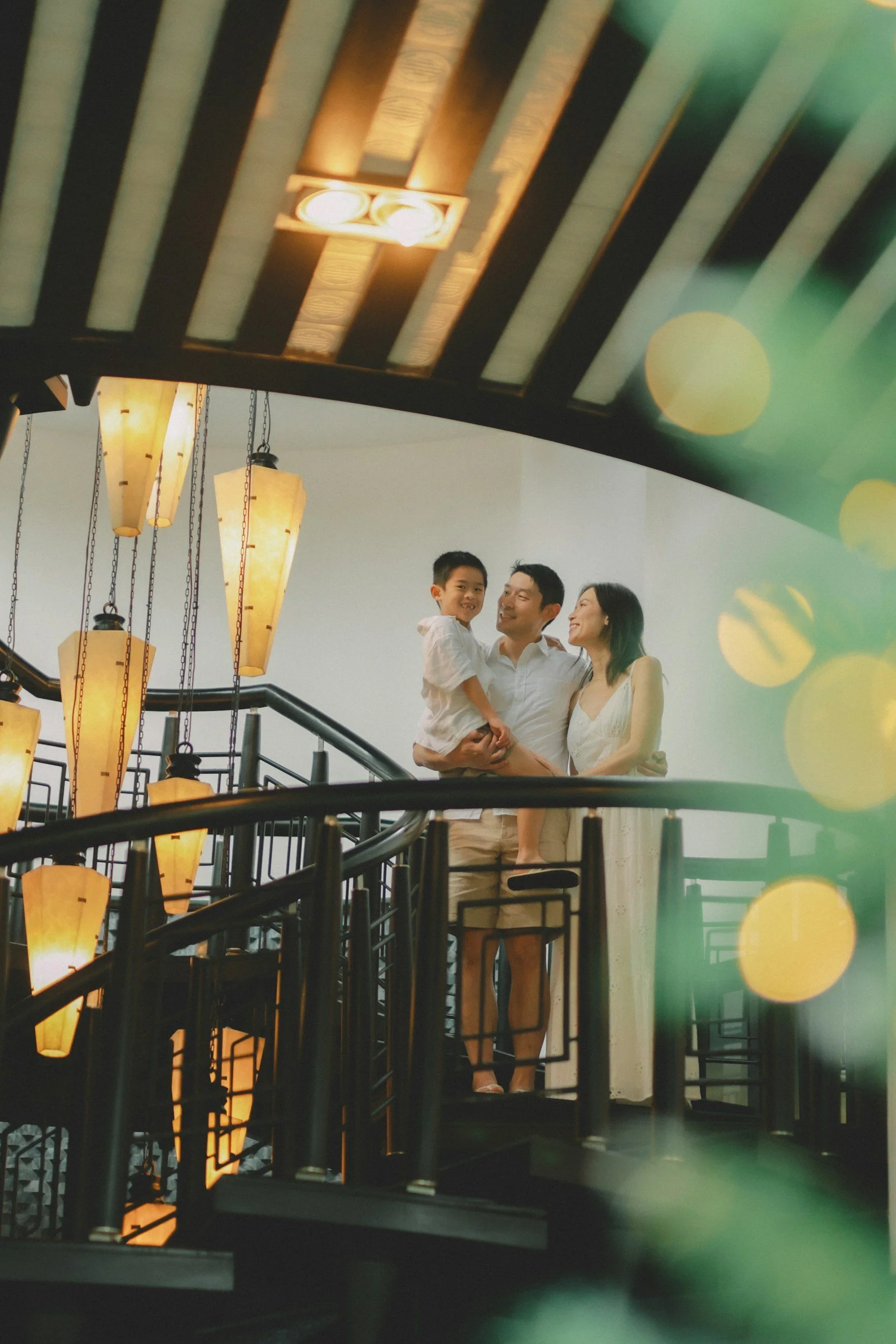 A family of three, with a young boy, their father, and mother, standing on a staircase, smiling and enjoying each other's company in a warmly lit interior space.