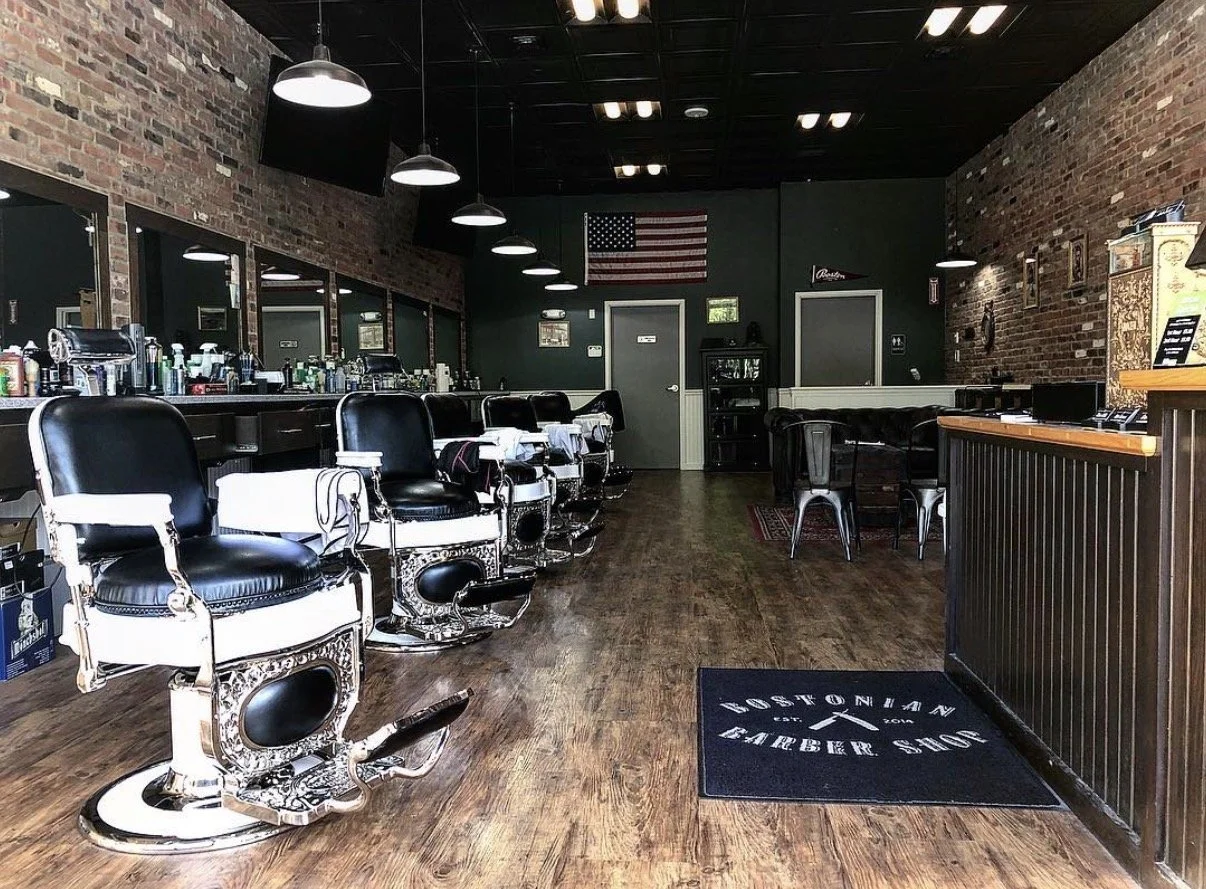 Interior of a barbershop with vintage barber chairs, hair products on a counter, a mirror wall, an American flag mural, and a reception desk.