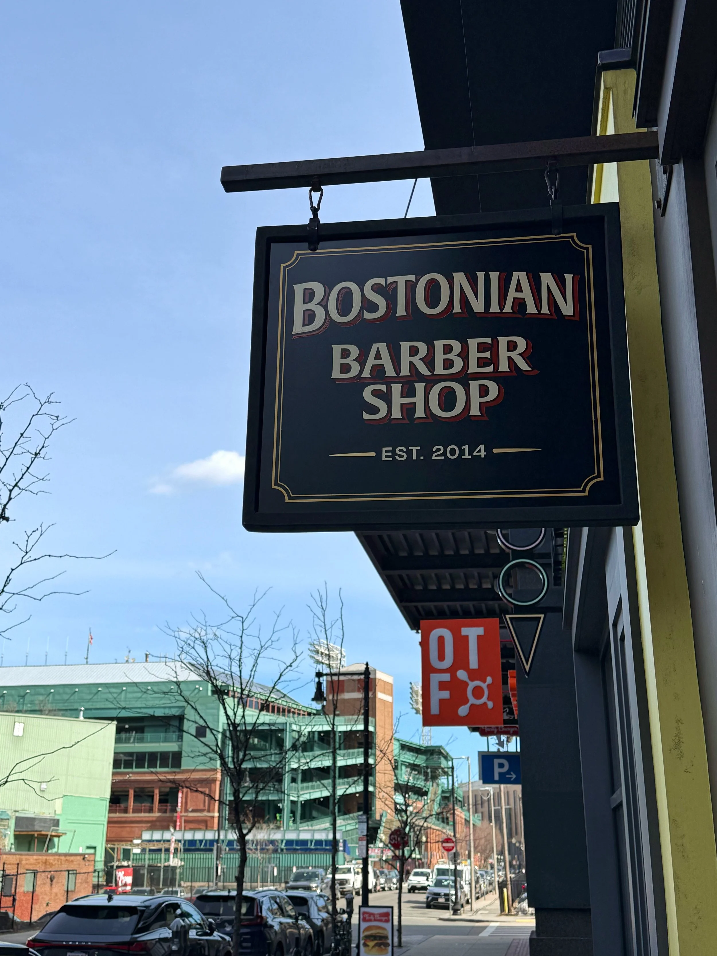 Street scene with signs including a large black sign for Bostonian Barber Shop established in 2014, a red and white 'TO' sign, and visible cars parked along the street with buildings and trees in the background.