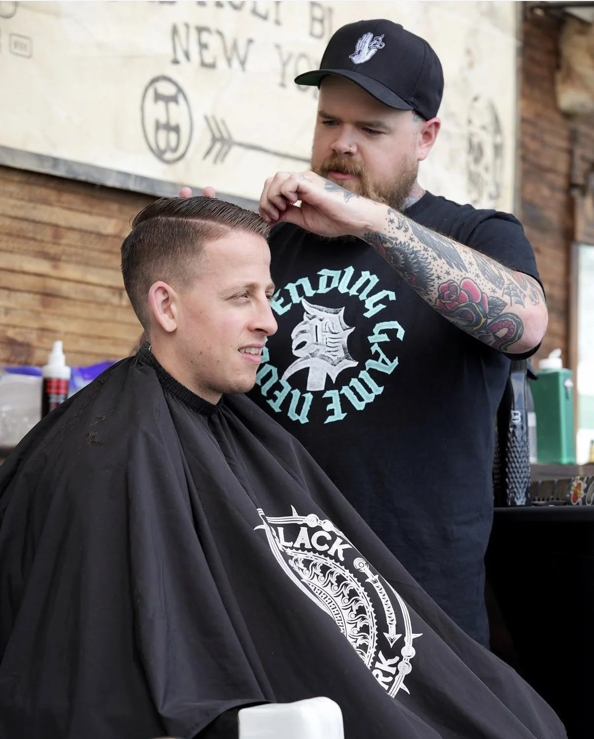 A barber wearing a black shirt and hat trims a man's hair in a barbershop. The man is seated with a black cape. The background features wooden walls and barber supplies.