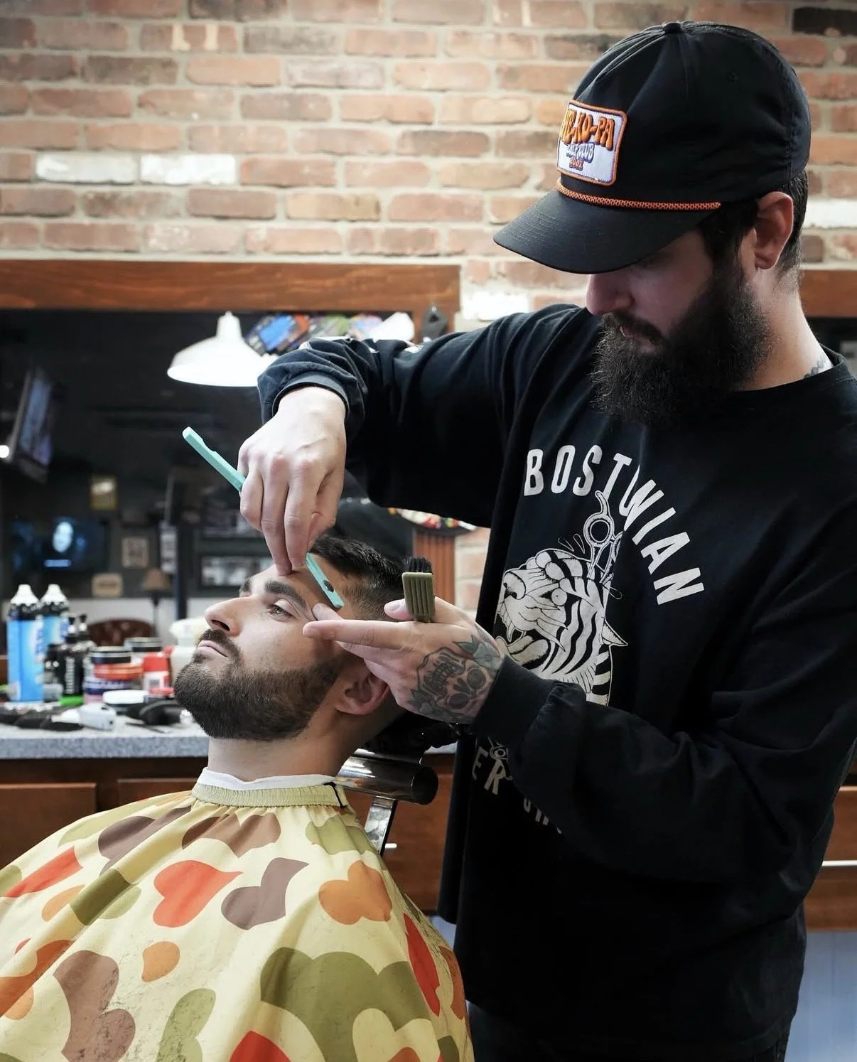 A barber is giving a close shave to a customer seated in a barber chair, with tools on the counter and a brick wall in the background.