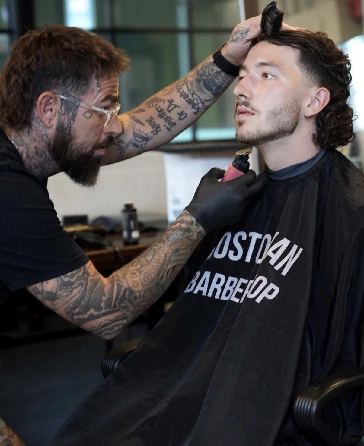 A tattooed barber is giving a haircut to a young man with dark, curly hair in a salon.
