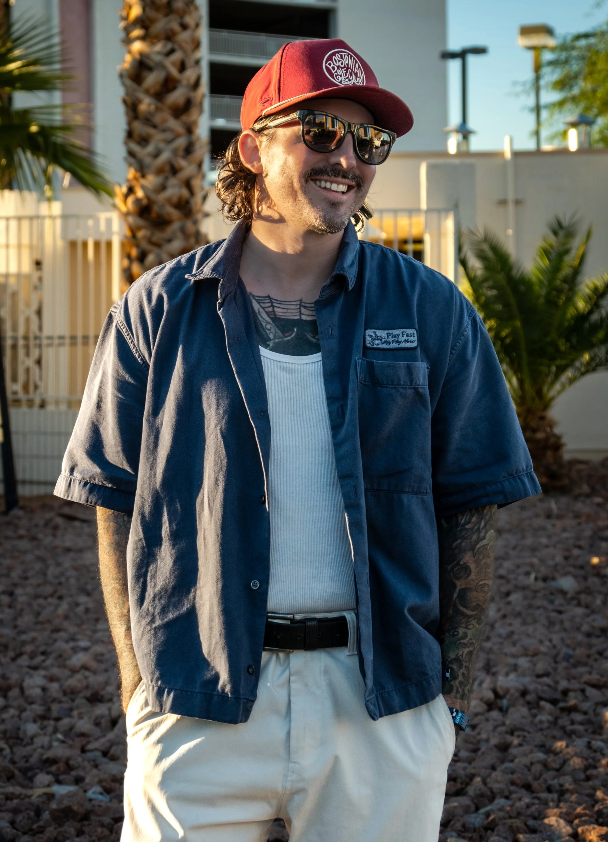 A man wearing sunglasses, a red cap, a blue short-sleeve button-up shirt, and beige pants stands outdoors in front of a building and trees during the daytime.