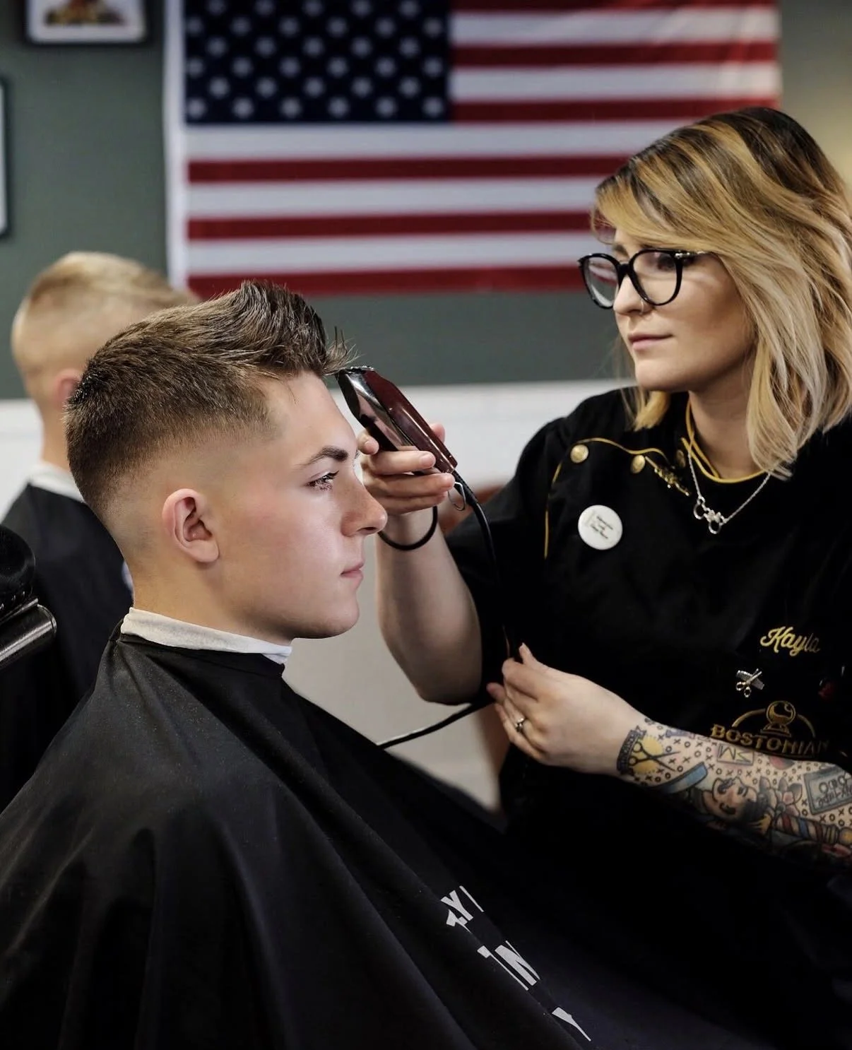 A barber applying makeup or face paint to a young man in a barbershop, with an American flag in the background.
