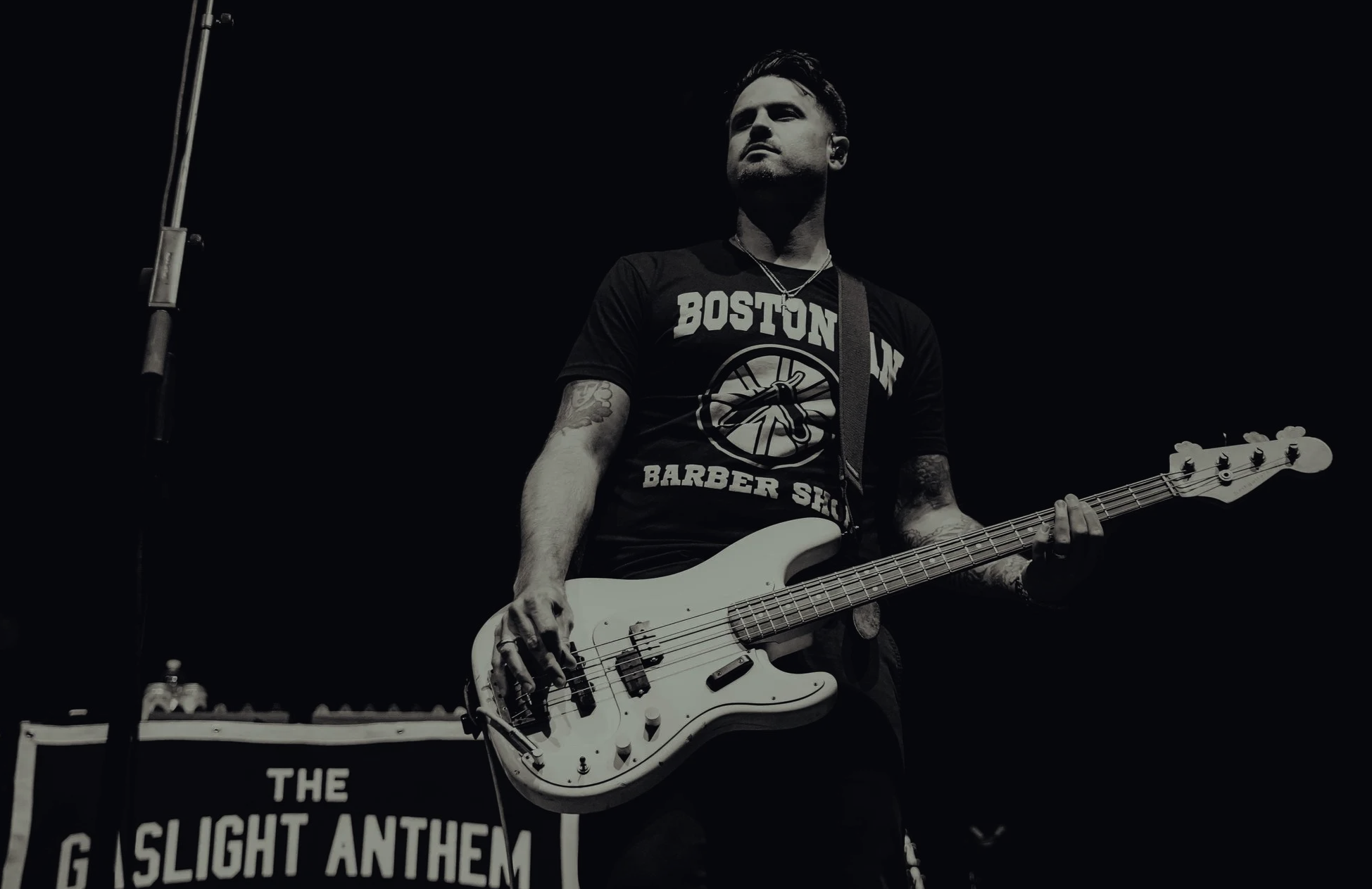 A male musician playing a bass guitar on stage, wearing a black T-shirt with ‘Boston’ and ‘Barber Shop’ written on it, with a microphone stand nearby and a banner reading ‘The Gaslight Anthem’ in the background.