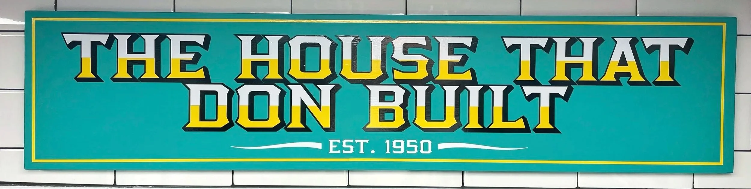 A sign that reads 'The House That Don Built, Est. 1950' in bold, black, white, and yellow letters on a teal background, bordered by a thin yellow frame.
