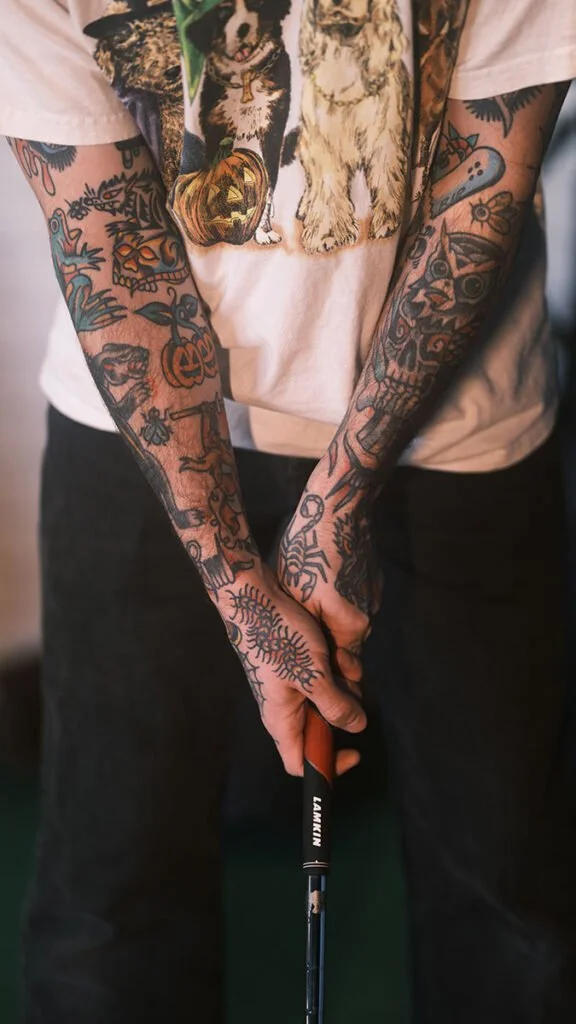 A person with extensive tattoos on both arms holding a golf club. The person is wearing a light-colored T-shirt with dog illustrations and black pants.