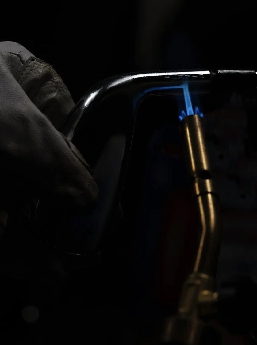 A person wearing gloves uses a blowtorch to weld or cut a piece of metal in a dark environment.