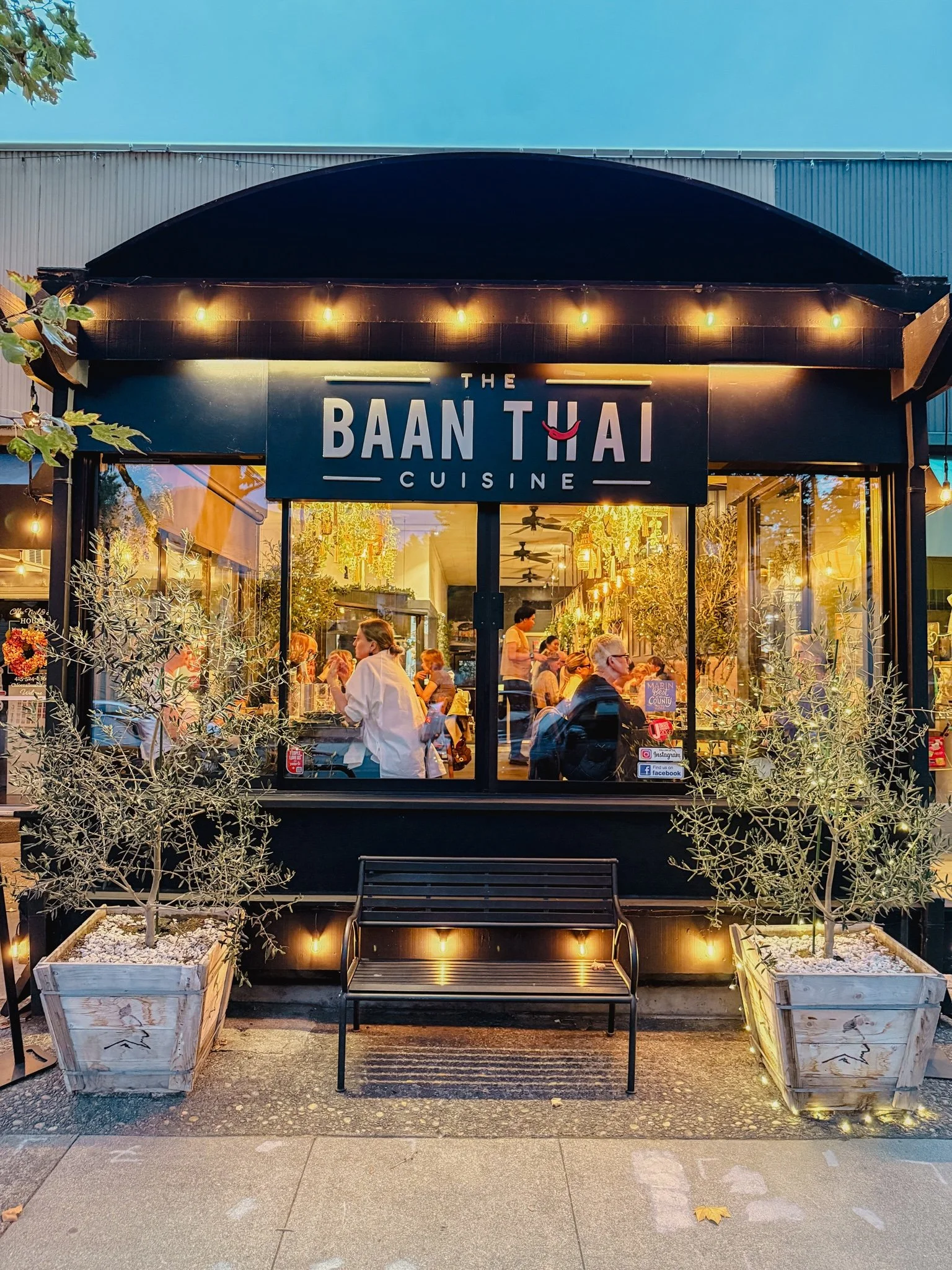 Front view of 'The Baan Thai Cuisine' restaurant with large glass windows revealing interior dining area, customers, and staff. Two potted trees are on either side of the entrance, illuminated by string lights and warm lighting.