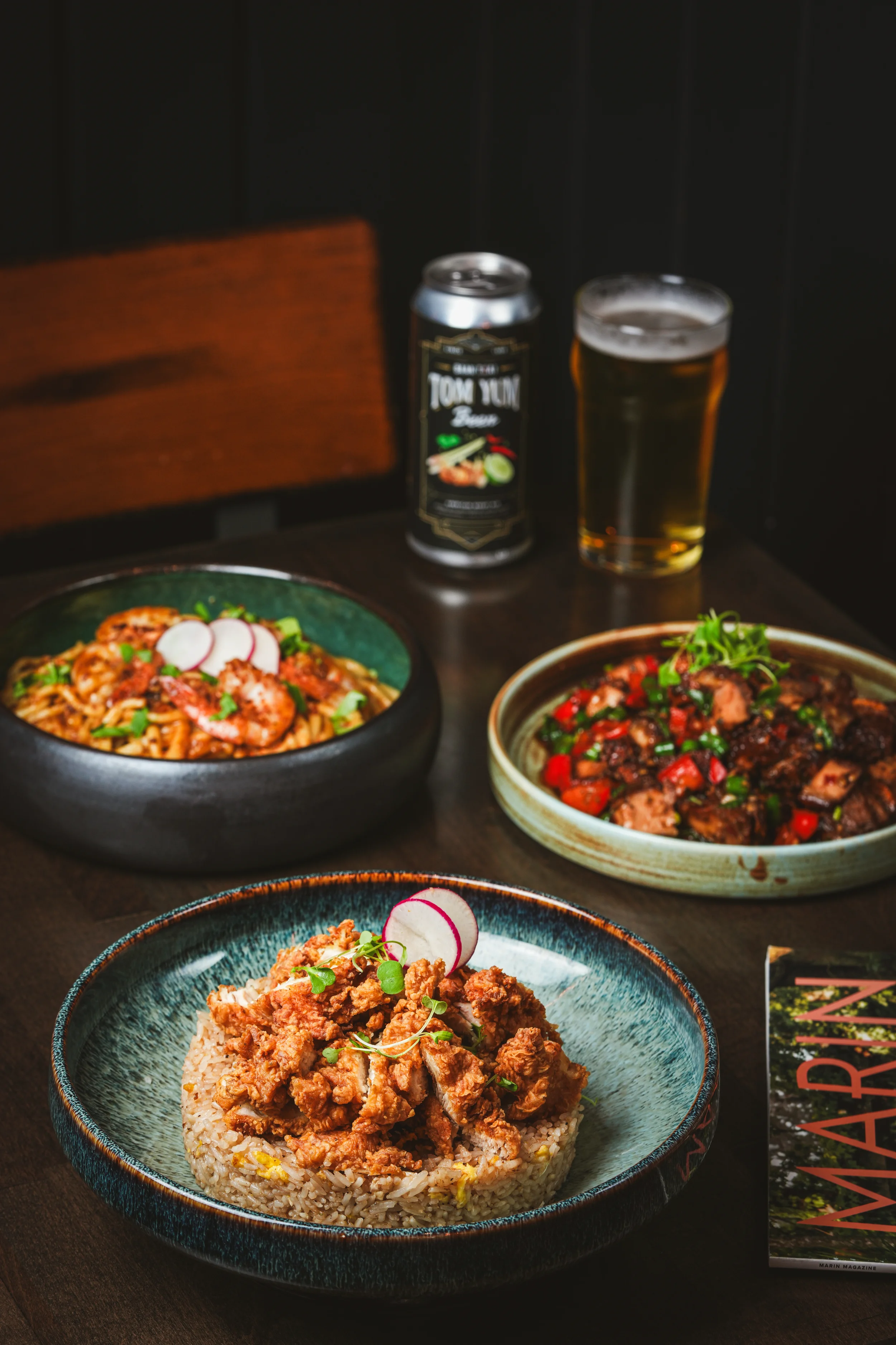 Several Asian dishes including fried chicken over rice, noodles, and stir-fried vegetables on a dark wooden table, with a can of Tom Yum beer and a glass of beer in the background.
