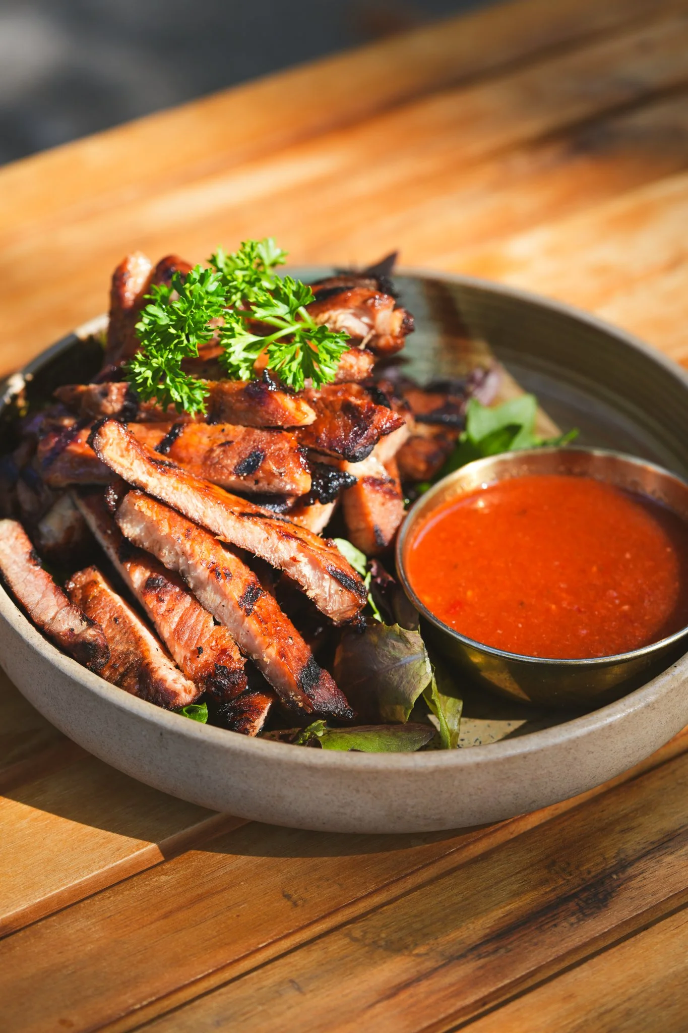 Grilled steak slices garnished with parsley, served with a side of red dipping sauce in a metal bowl on a gray ceramic plate, on a wooden table.