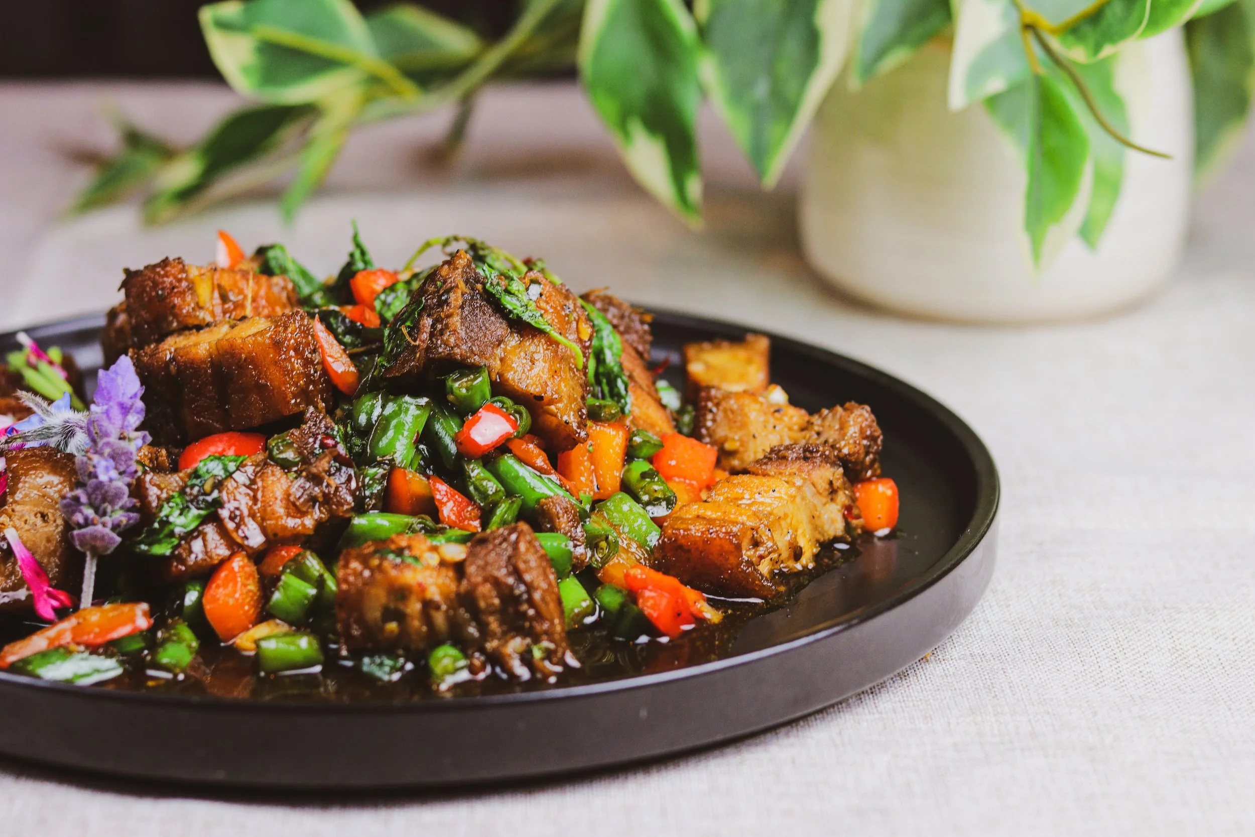 Cooked meat with vegetables on a black plate, garnished with purple flowers, in front of a plant in a white pot.
