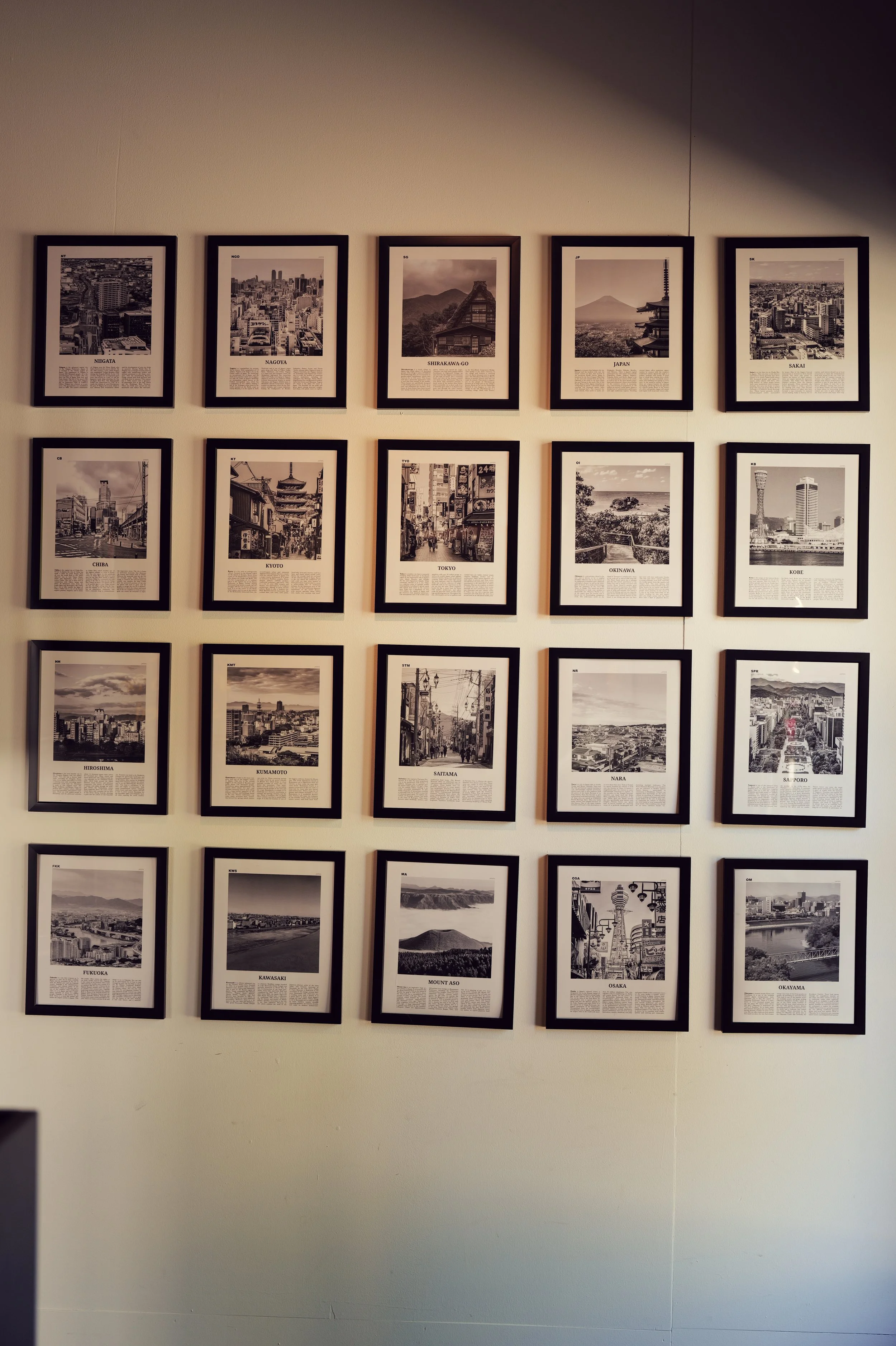 A wall display of 20 framed photographs of various cities in Japan arranged in a grid pattern with four rows and five columns. Each photo has a caption with the city name and a description.