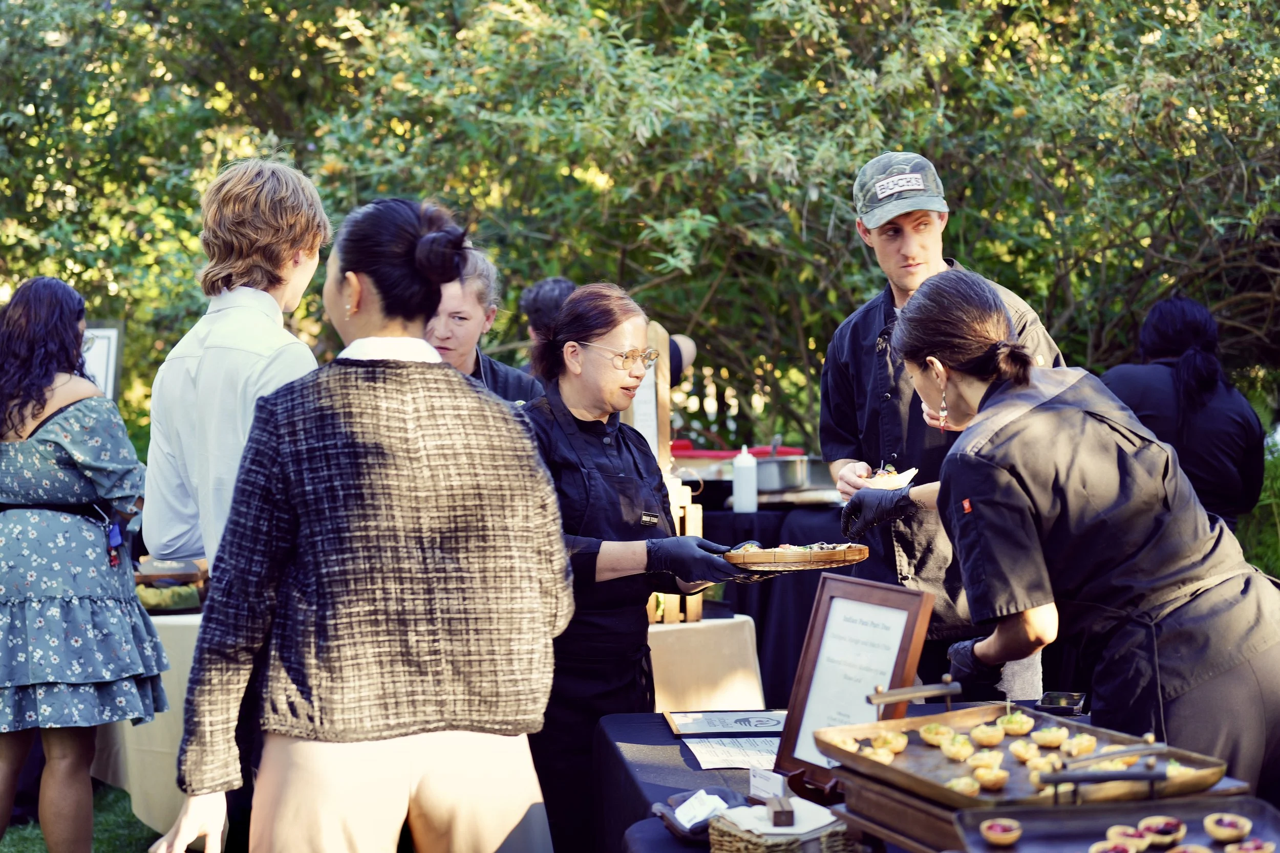 People at an outdoor event around a food table, with some ordering and others preparing or serving food, set against green trees and sunny weather.