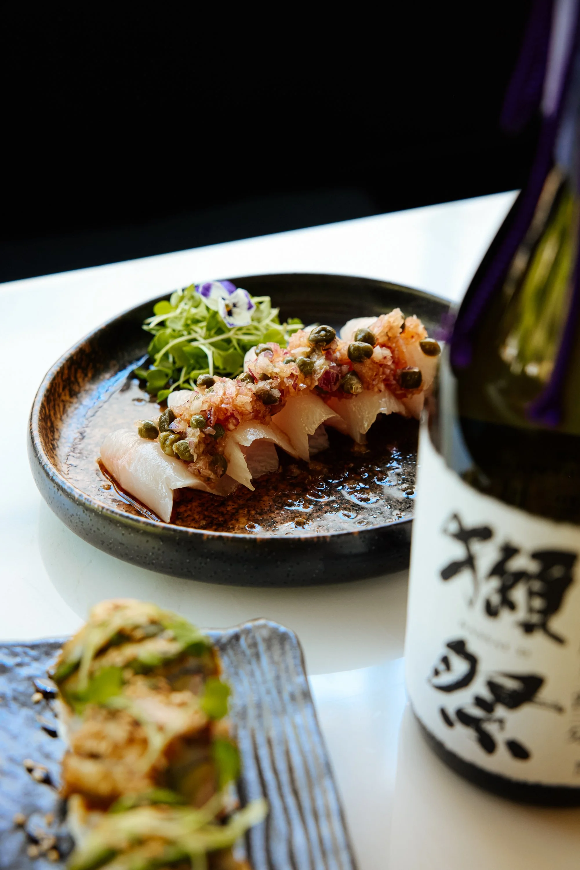 A black ceramic plate with slices of sashimi topped with chopped green herbs and a side of microgreens, with a bottle of sake labeled in Japanese in the background.
