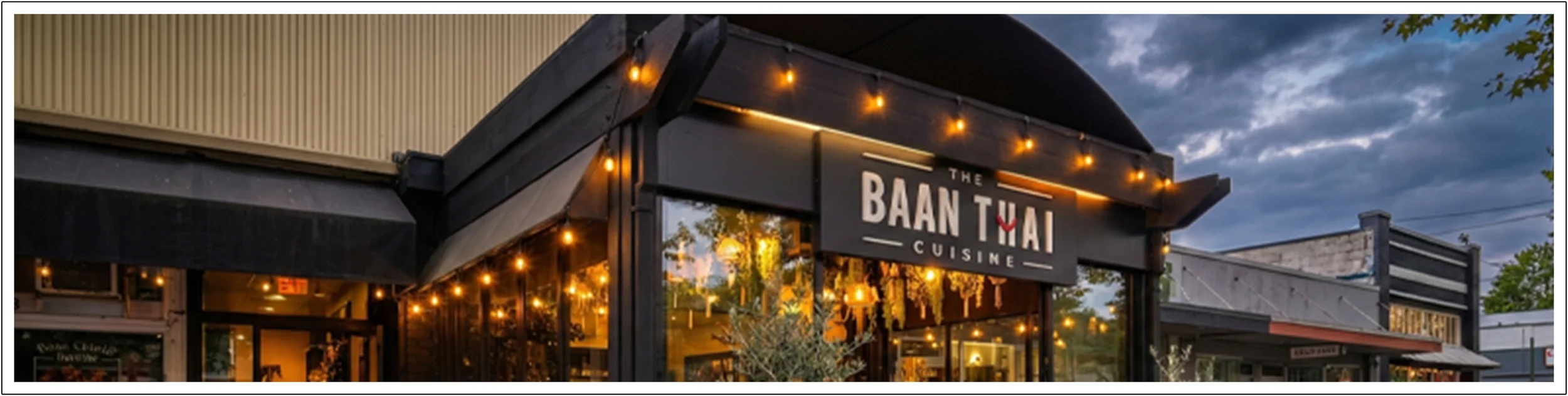 Exterior of a restaurant called The Baan Thai Cuisine with string lights and large windows during evening with cloudy sky.