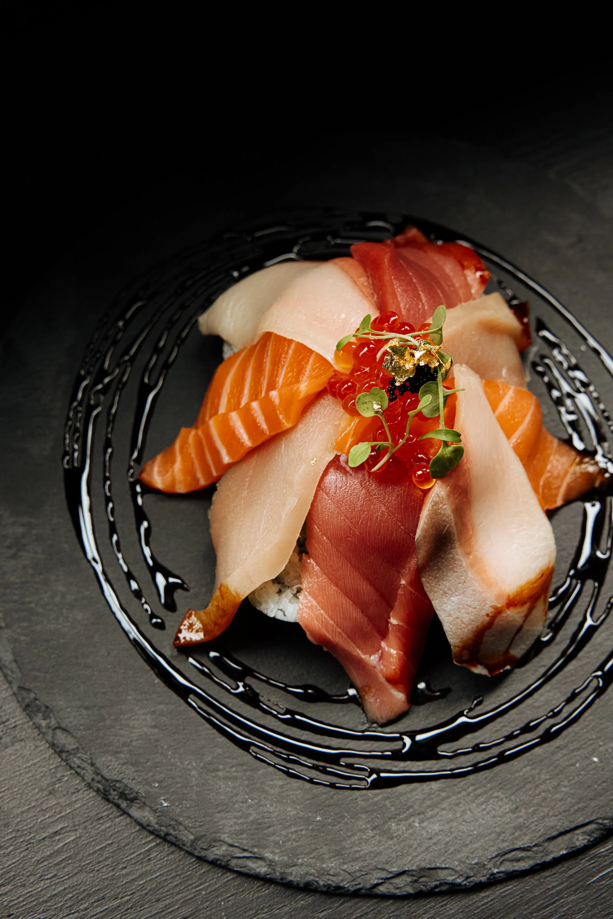 Assorted slices of raw fish, including salmon, tuna, and white fish, arranged on a black slate plate decorated with a circular drizzle of sauce and garnished with microgreens and fish roe.