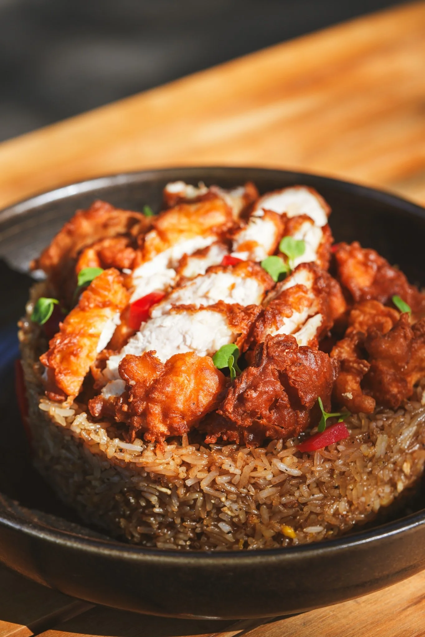 A bowl of Asian-style fried rice topped with sliced fried chicken, crispy fried chicken pieces, and garnished with small green herbs.