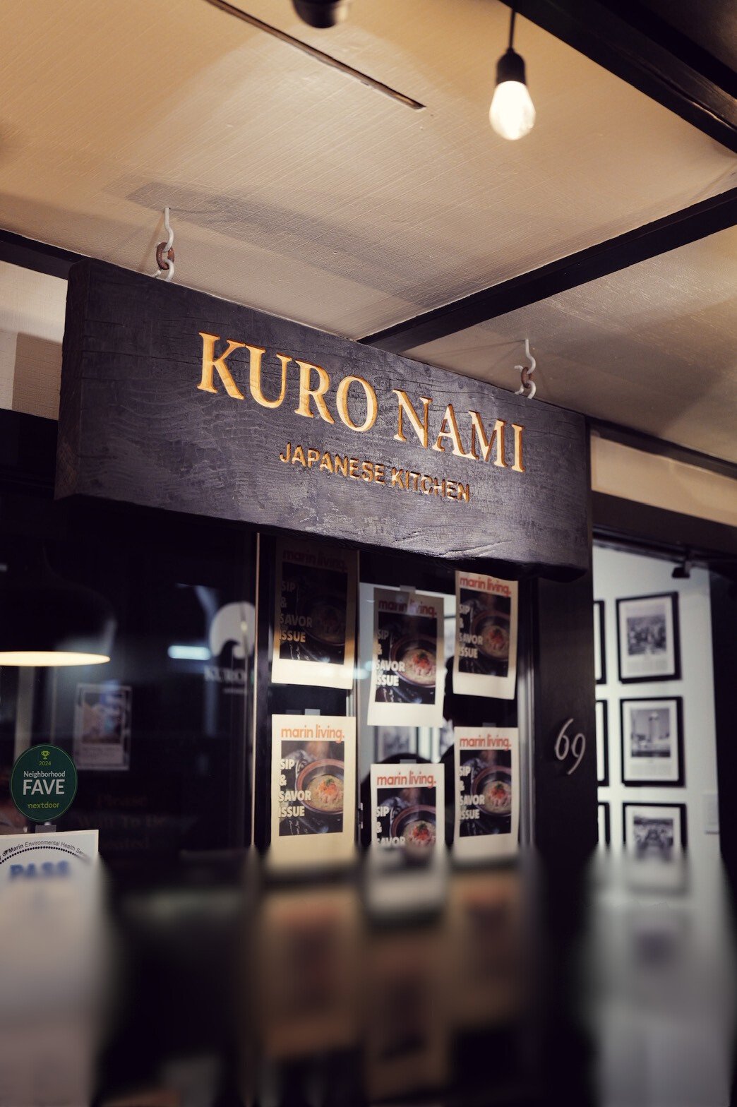 A restaurant sign reading "KURO NAM" with a subtitle "JAPANESE KITCHEN" hanging from the ceiling in a restaurant interior, with photos and a green sticker on a glass door or window below.