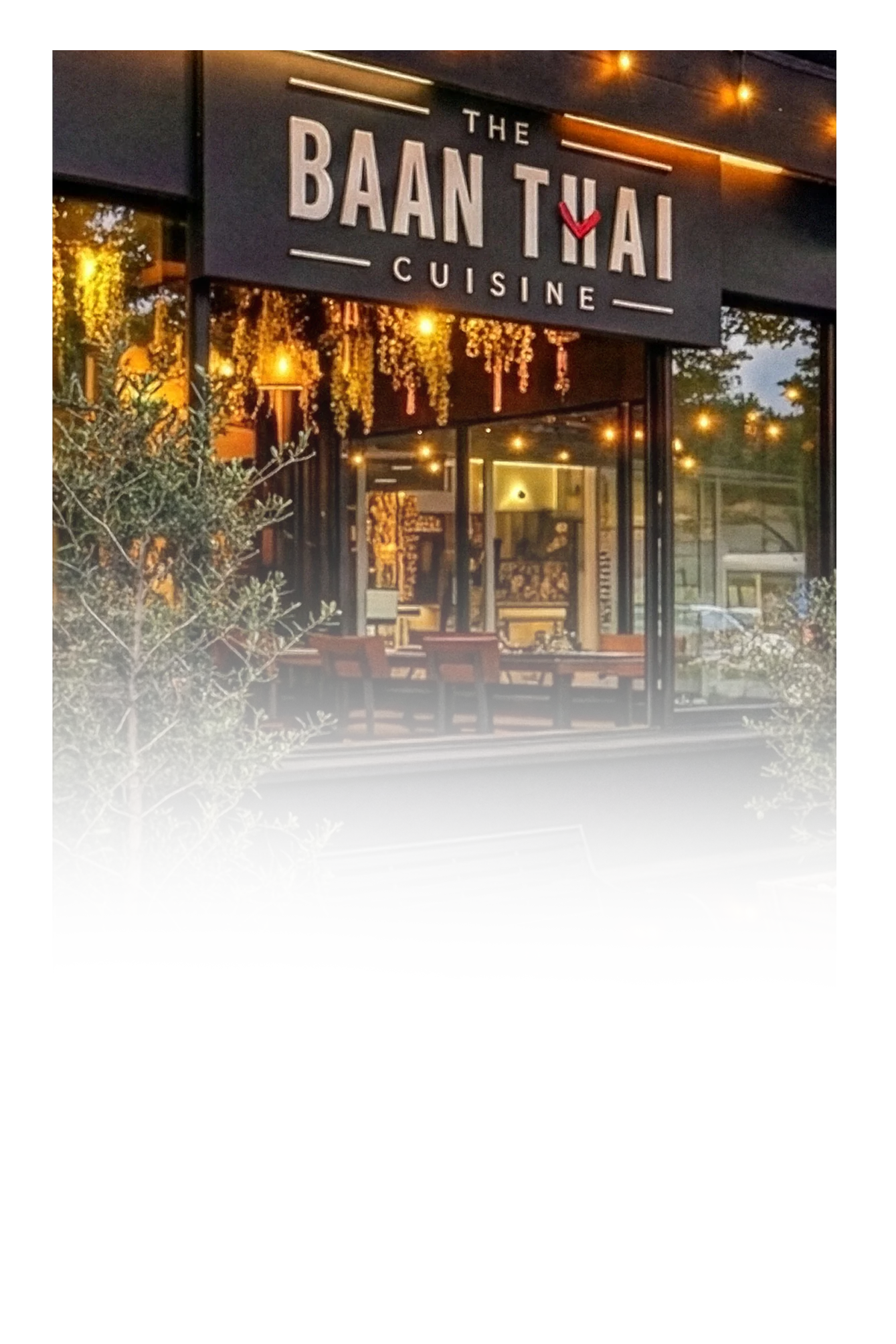 Exterior view of a restaurant named 'The Baan Thai Cuisine' with a large sign, warm interior lighting, and outdoor seating area surrounded by trees.