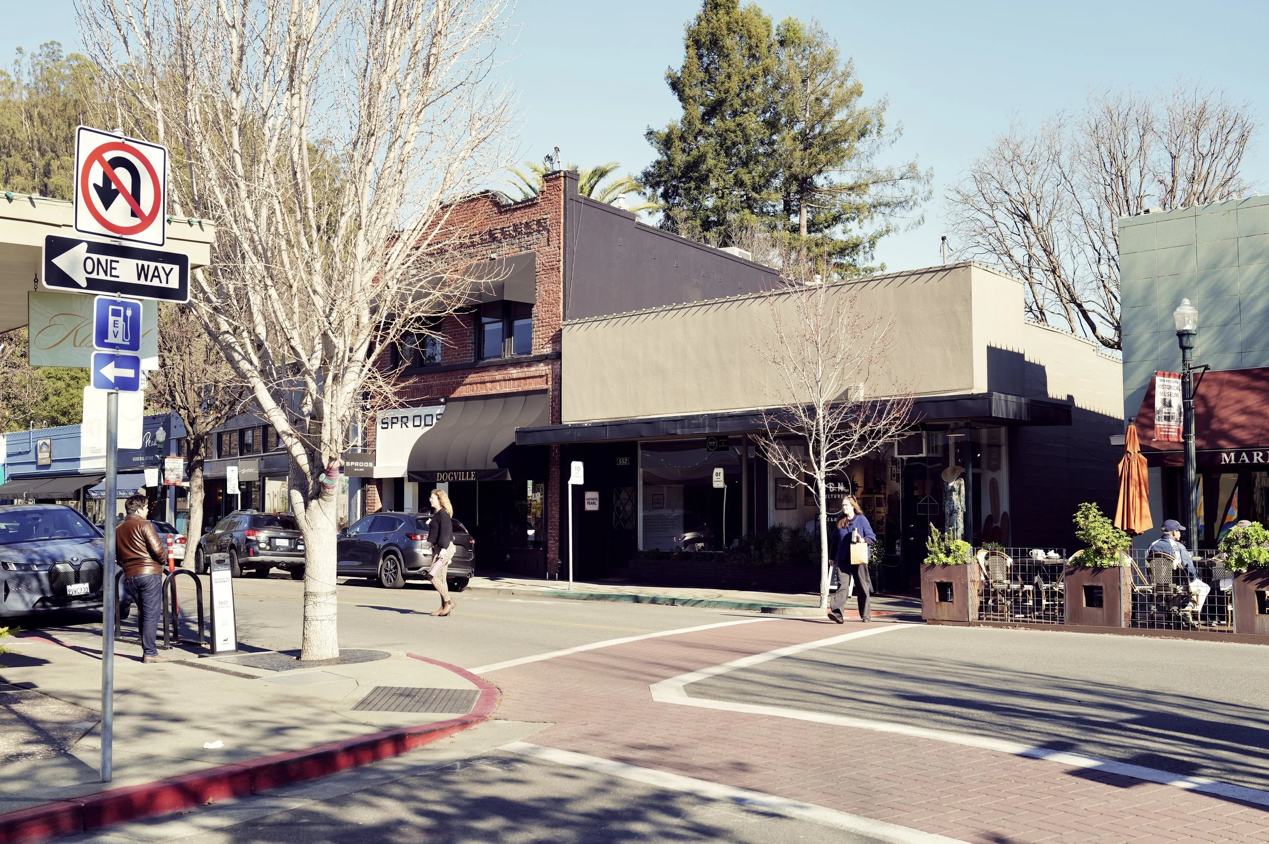 Downtown street scene with storefronts, parked cars, pedestrians, leafless trees, and street signs in clear weather.