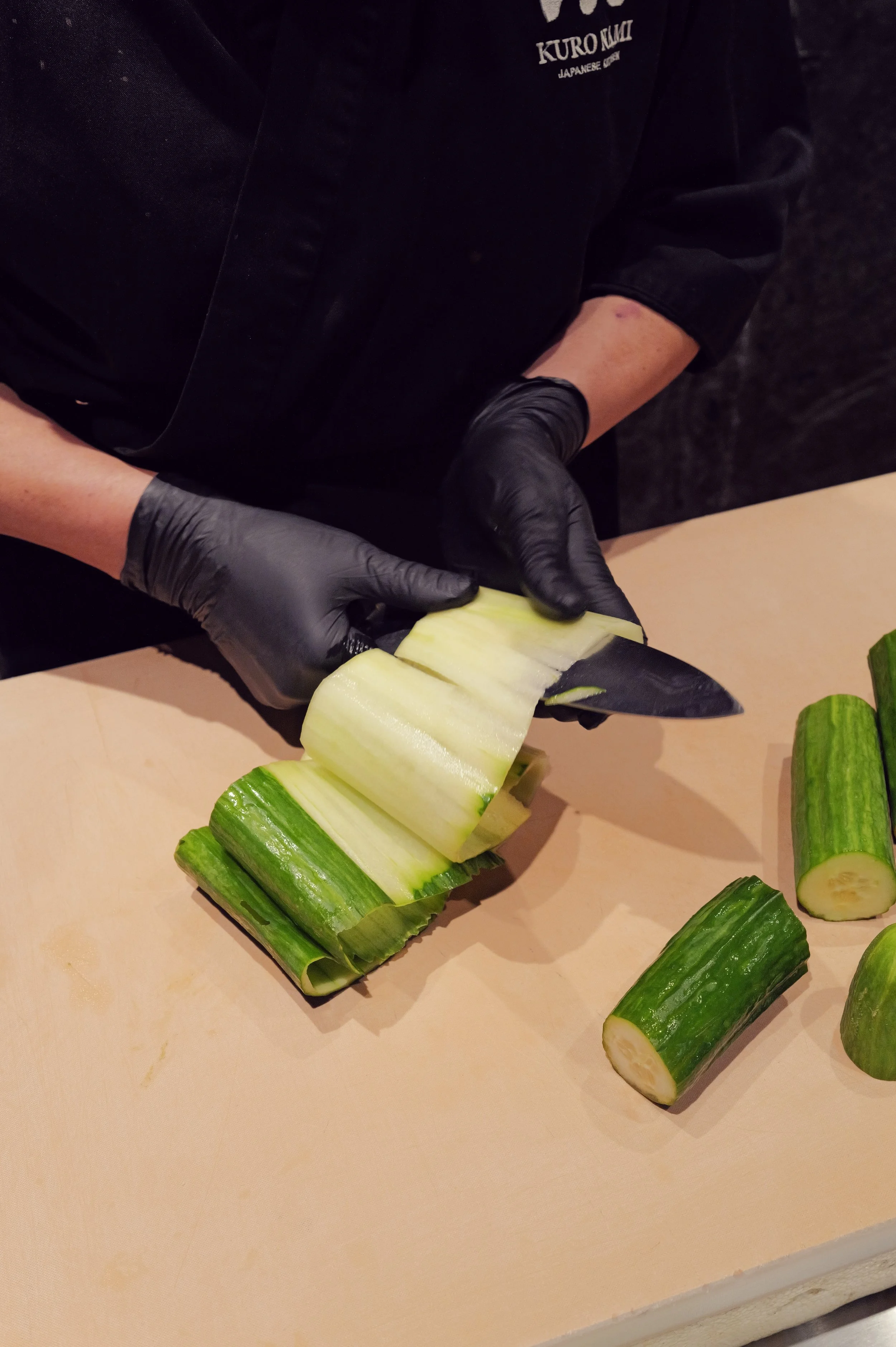 Person wearing black gloves and a black uniform slicing a cucumber on a wooden cutting board.