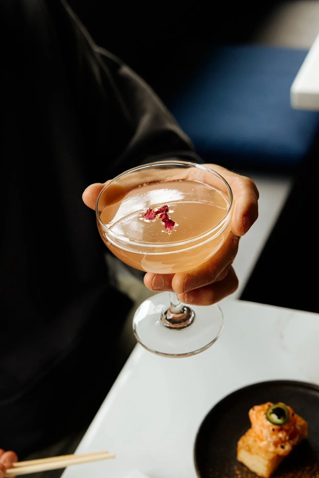 A hand holding a coupe glass filled with a pink cocktail garnished with dried flower petals, with a black plate of food nearby on a white table.