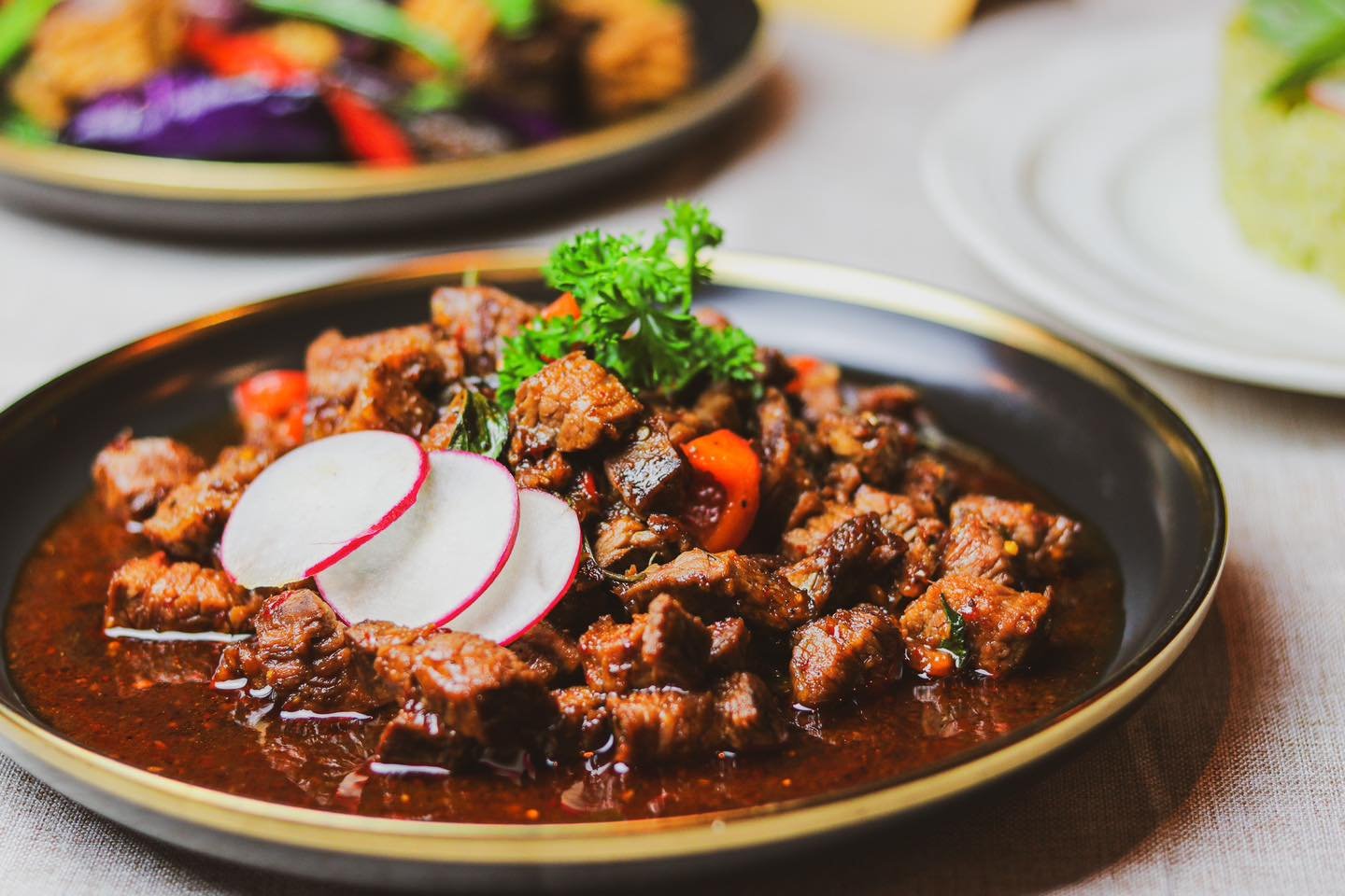 A black plate with beef stew garnished with fresh parsley and garnished with slices of radish, with other dishes visible on a light tablecloth.