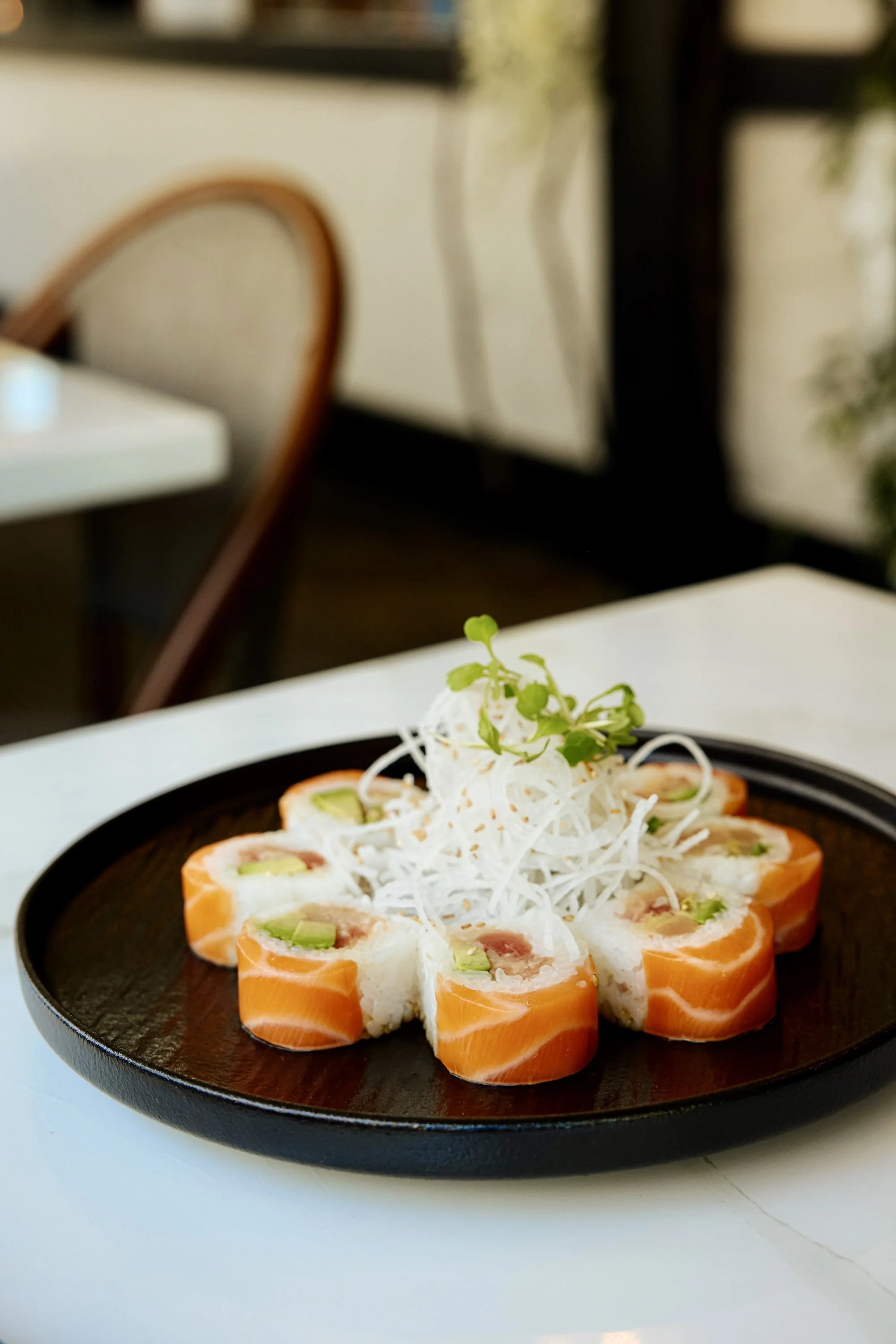 A black plate of salmon sushi rolls garnished with shredded daikon and microgreens on a white table in a restaurant.