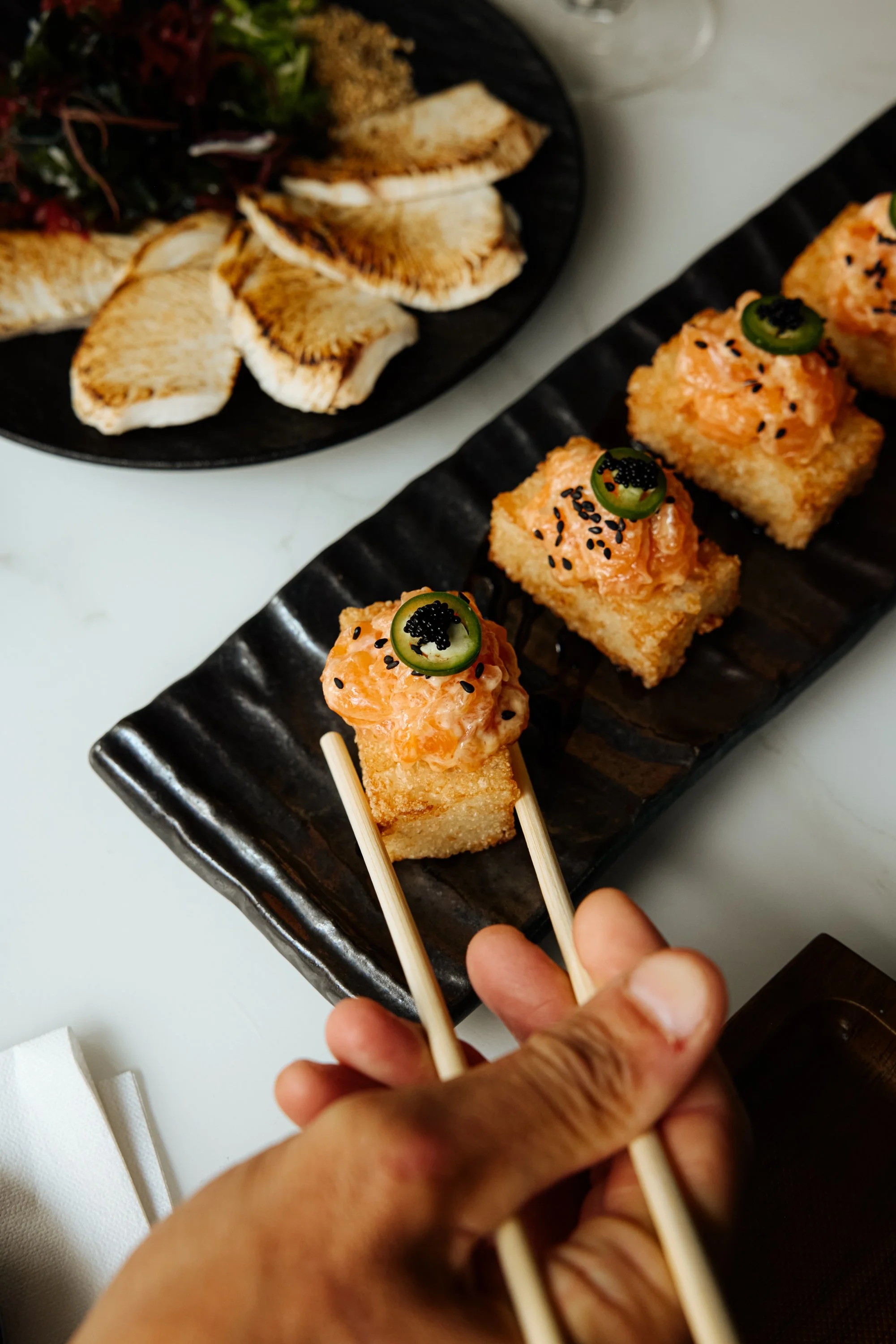 Close-up of a hand holding chopsticks with a piece of sushi topped with orange fish, green pepper slice, black sesame seeds, and black fish roe, on a black rectangular serving plate. In the background, there is a black plate with grilled halibut steak and salad on a white surface.