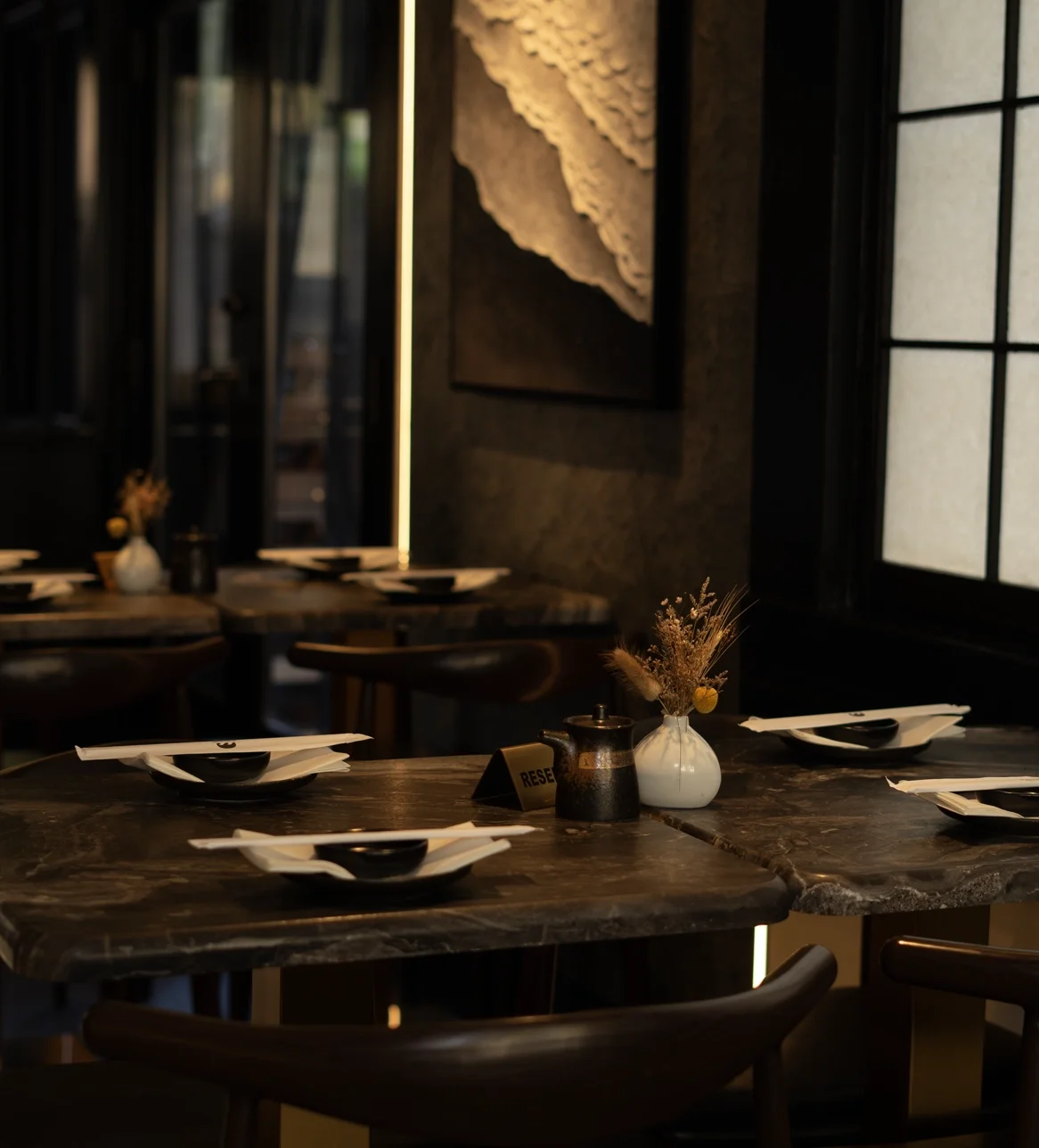 Empty restaurant table set with black and white dishes, small vase with dried flowers, and a reserve sign in a dimly lit, modern dining space with dark decor and large windows.