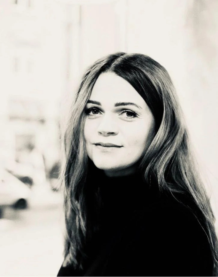 Schwarz-weiße Aufnahme einer jungen Frau mit langem, leicht gewelltem Haar, die in die Kamera schaut, vor einem unscharfen Hintergrund.