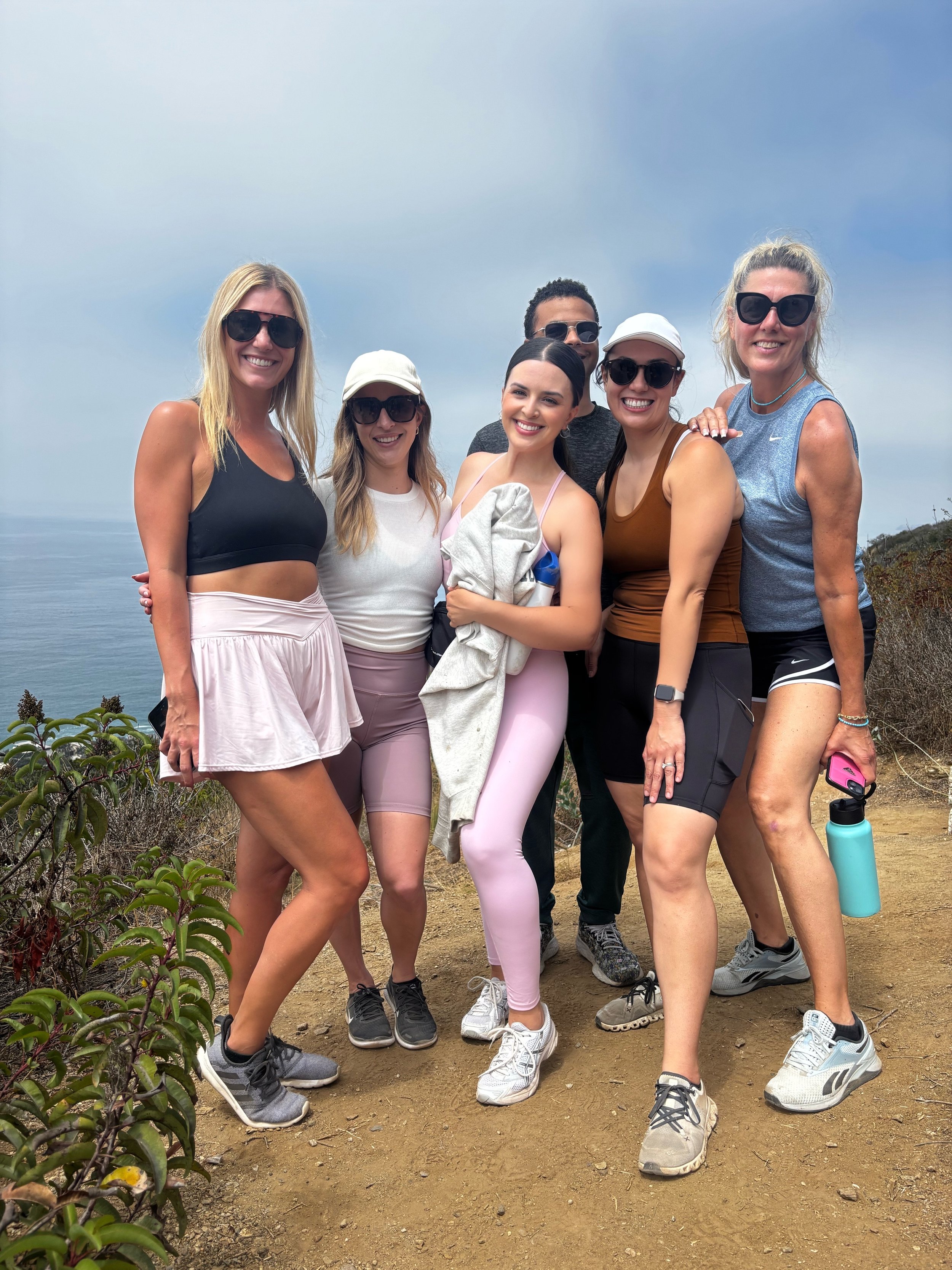 Group of six women and one man standing on a hiking trail outdoors, with ocean and cloudy sky in the background.