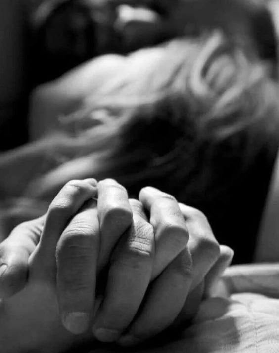 Close-up of two people holding hands, with a blurred person lying in the background.