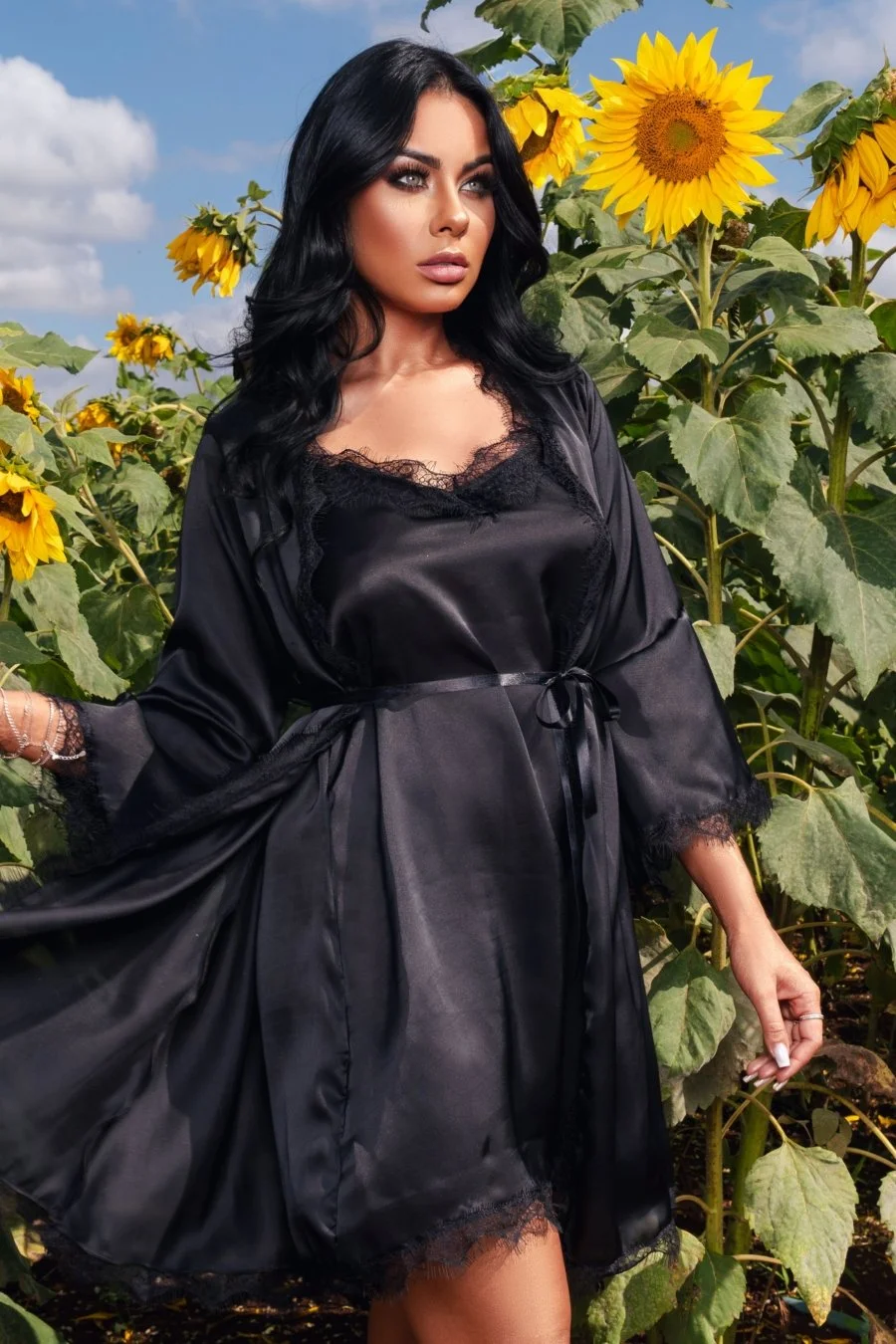 A woman with long black hair and makeup wearing a black satin robe with lace trim, standing among tall sunflowers outdoors during daytime.