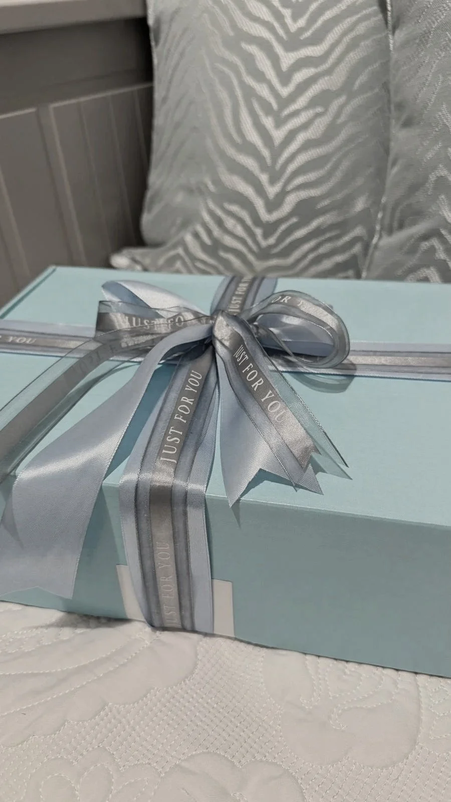 Light blue gift box wrapped with gray ribbon that says 'Just for you', placed on a white tablecloth with a quilted floral pattern.