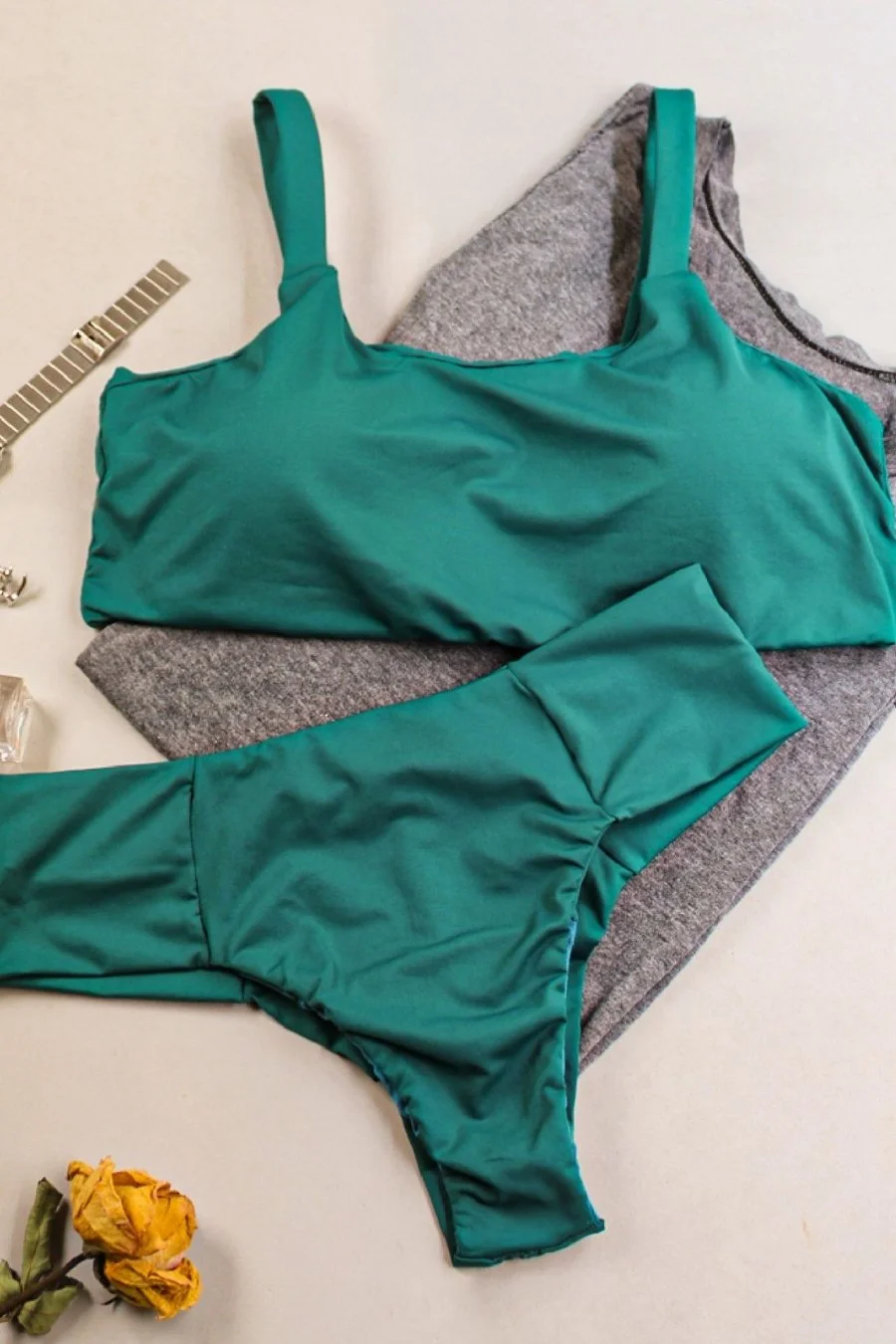 A teal bikini laid out on a gray cloth, with a gray and black garment nearby, on a white surface, with a yellow dried flower and a watch with a metal band and a ring also visible.