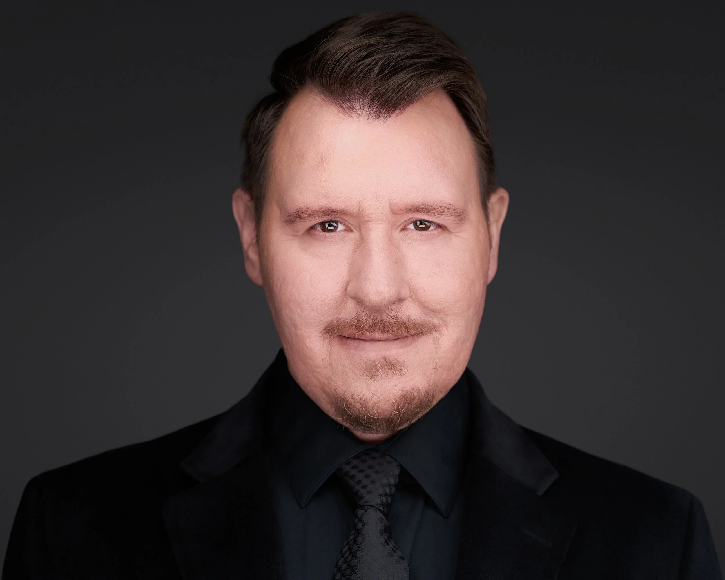 Close-up portrait of a man with short, dark hair and a beard, dressed in a black suit and tie, against a solid dark background.