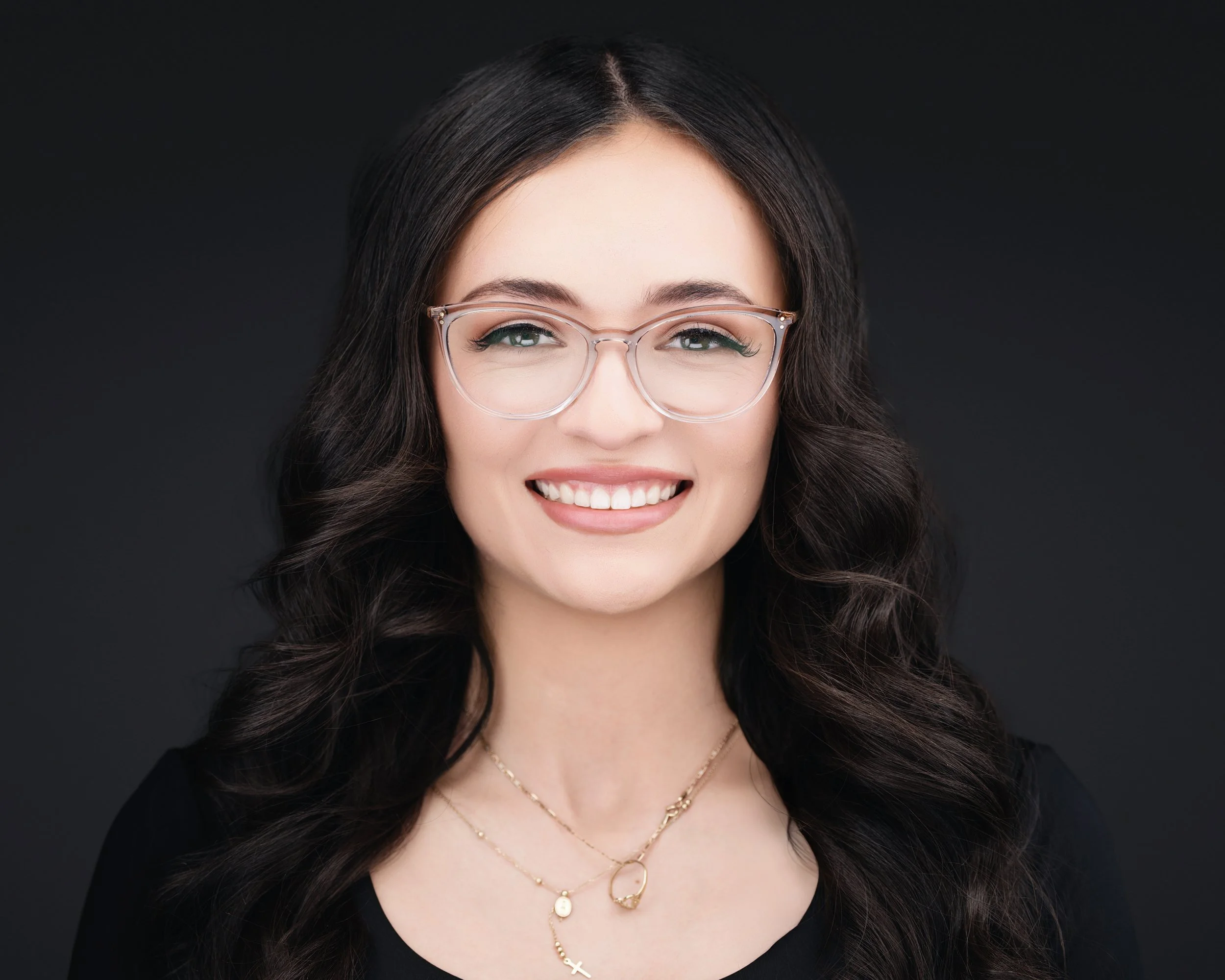 A smiling woman with dark wavy hair, wearing glasses, gold layered necklaces, and a black top, against a dark background.