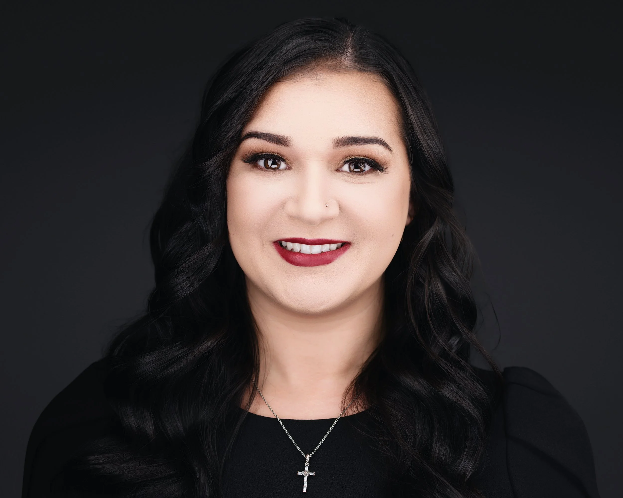 Close-up of a woman with black hair, brown eyes, makeup, dark red lipstick, wearing a black top and a silver cross necklace, against a dark background.