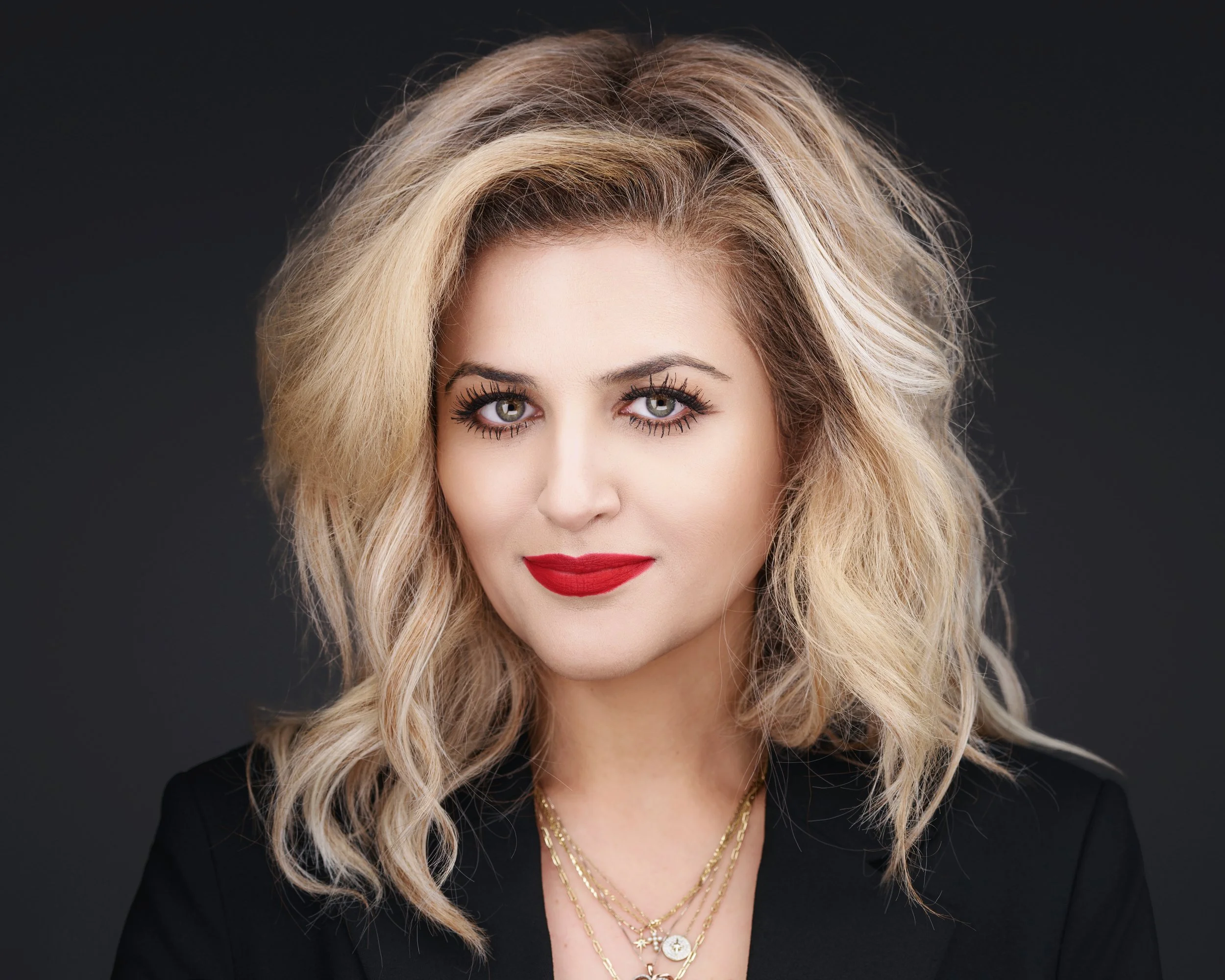 A woman with wavy blonde hair, wearing red lipstick, black top, and layered gold necklaces, against a dark background.