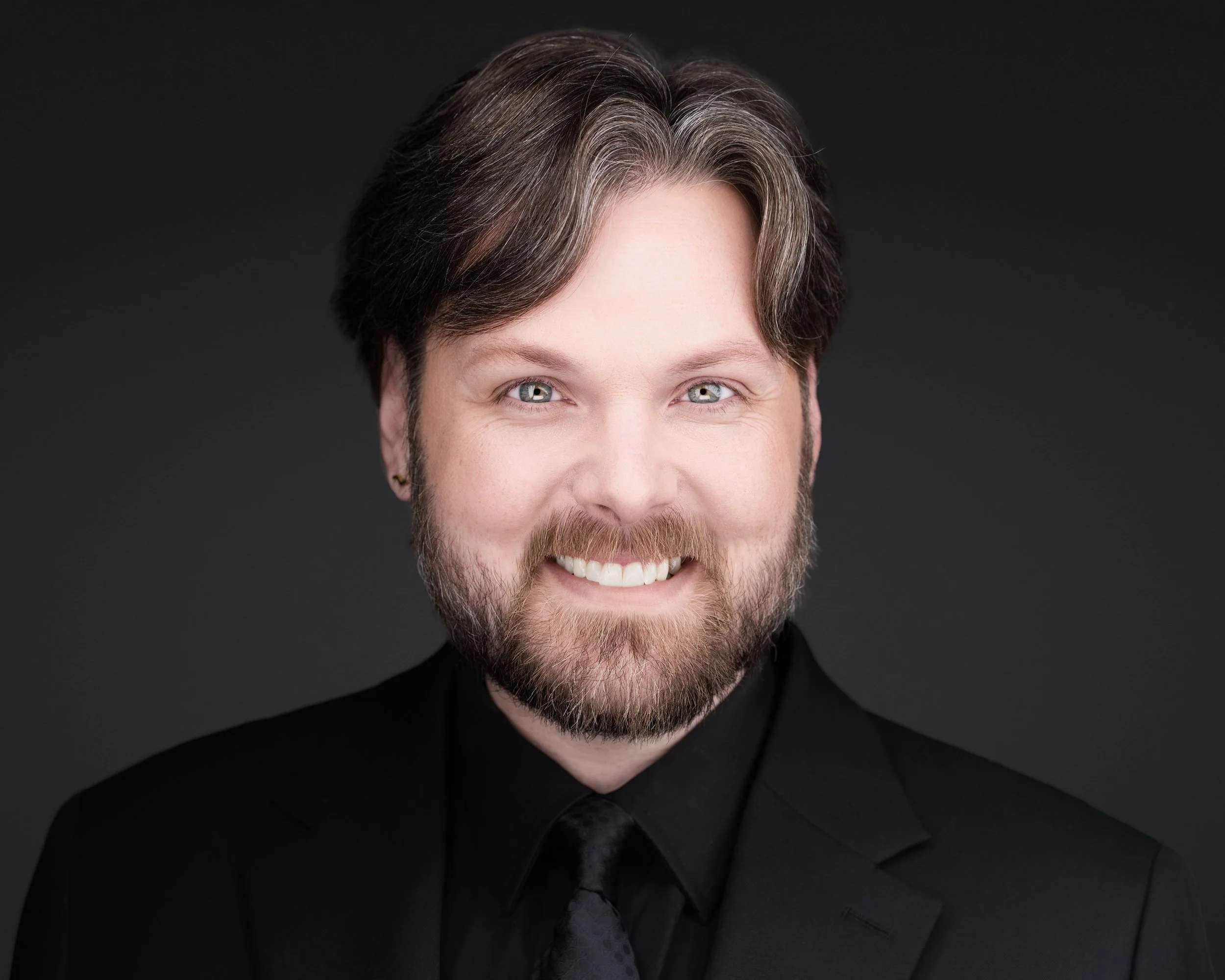 Close-up of smiling man with gray and brown hair, beard, and blue eyes, wearing a black suit jacket against a dark background.