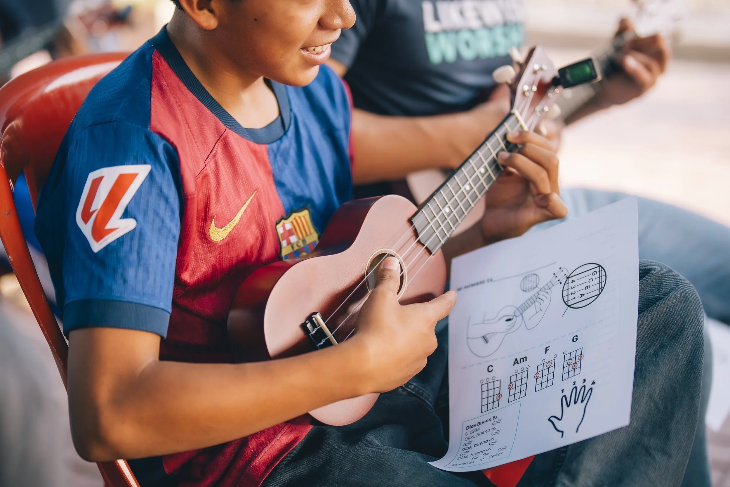 A young boy wearing a Barcelona soccer jersey playing a ukulele while holding sheet music that has guitar chords and a drawing of a guitar.