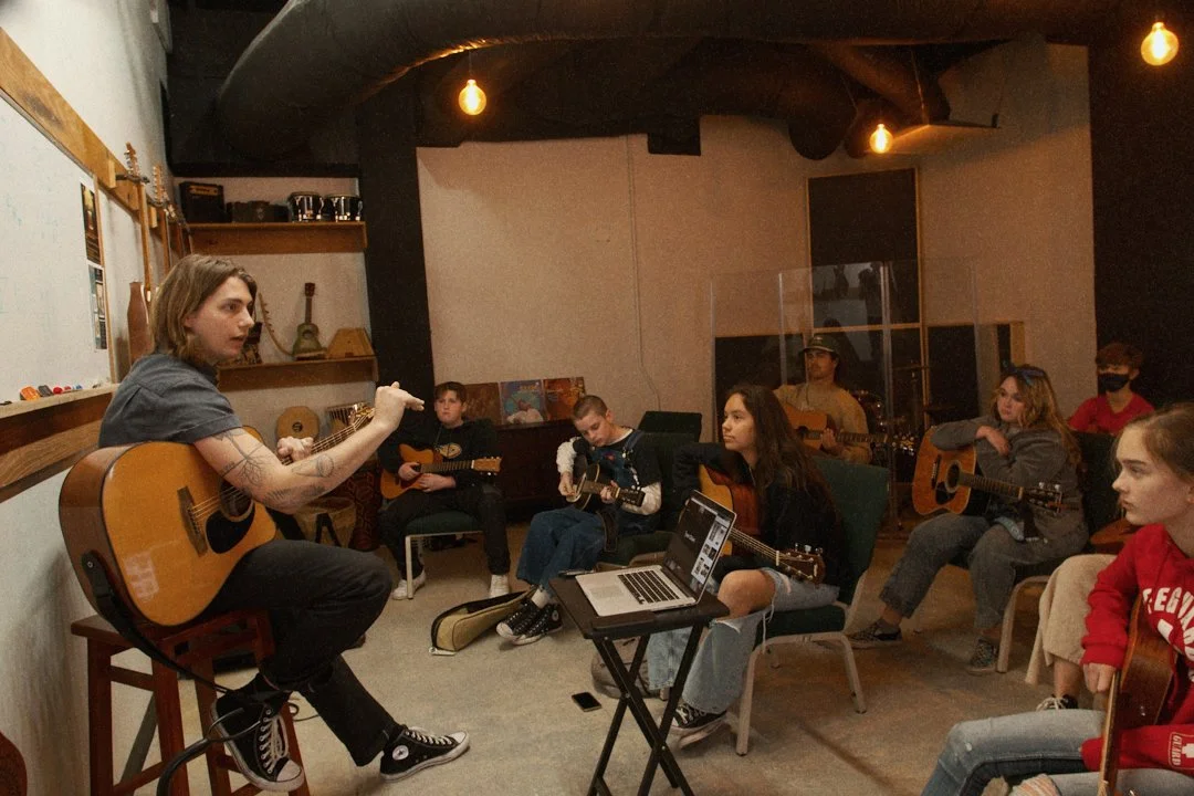 Young woman instructing a group of children and teenagers in a music lesson, all playing acoustic guitars, in a cozy indoor room with warm lighting.