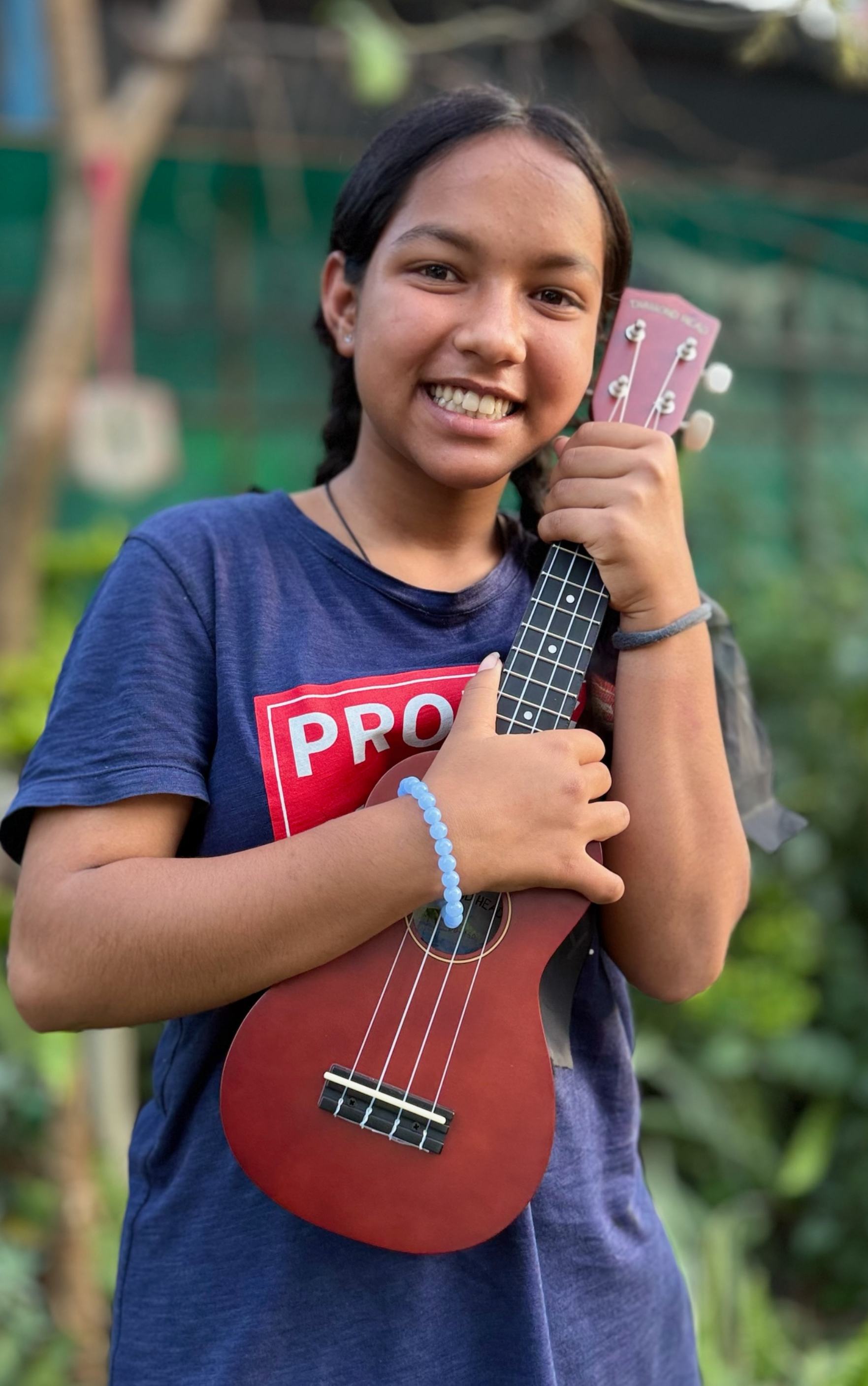 A young girl smiling outdoors holding a small red ukulele close to her chest, wearing a navy blue T-shirt and a blue bracelet, with a green blurred background.
