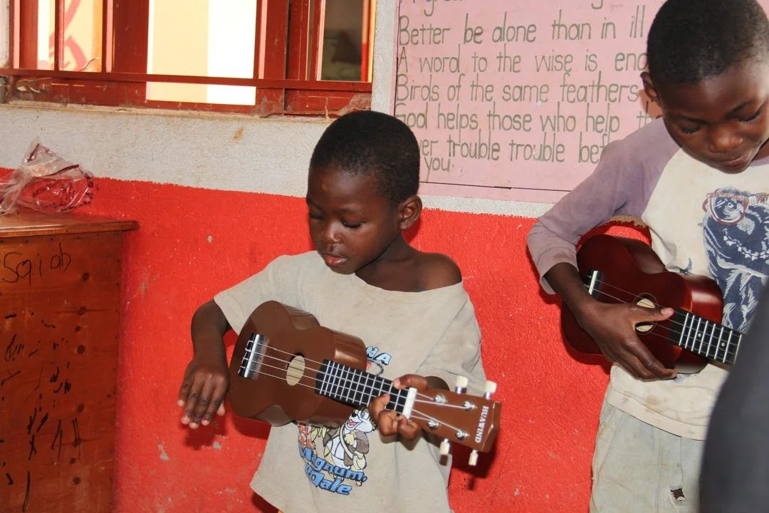 Two young boys playing small brown ukuleles inside a classroom with red and white walls.